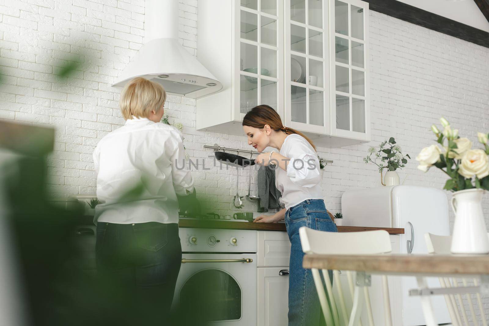 The older mother prepares food in the kitchen, the daughter drinks tea and looks at her mother.