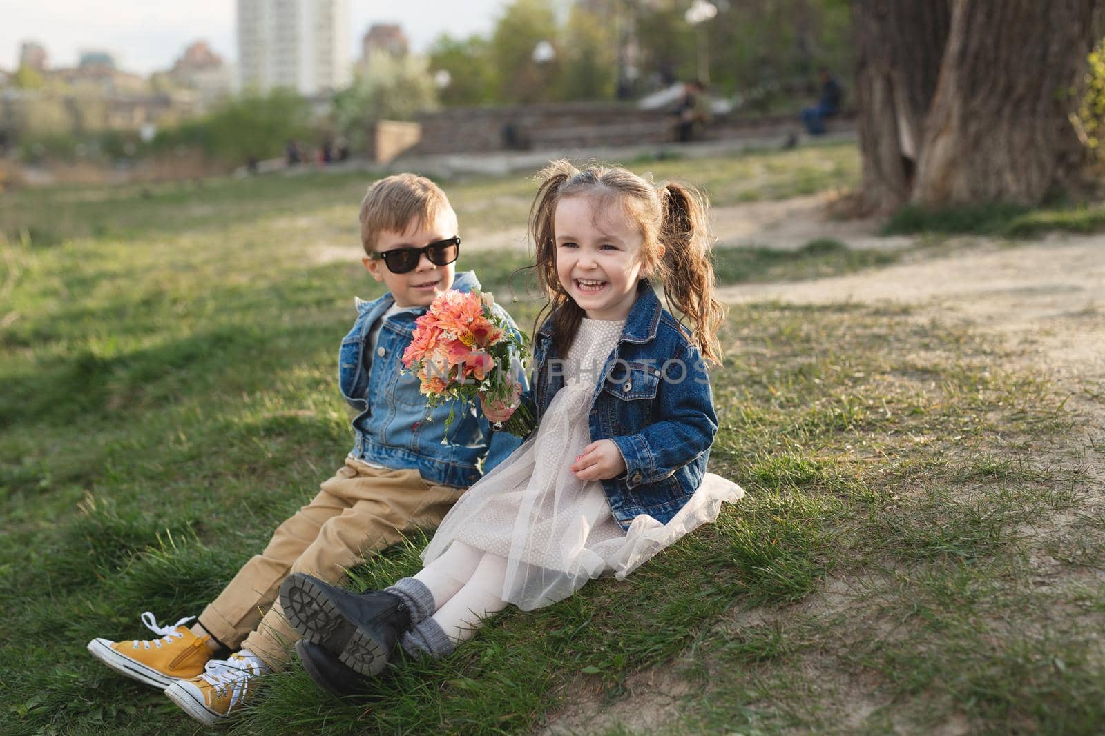 A little happy cute boy and a little cute girl are sitting on the grass in a beautiful place with a bouquet of flowers.