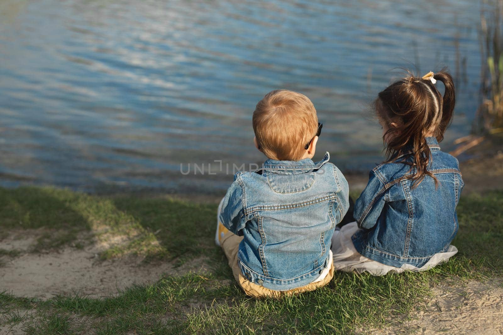 Back view of a little boy and girl sitting on the shore of a pond at sunset.
