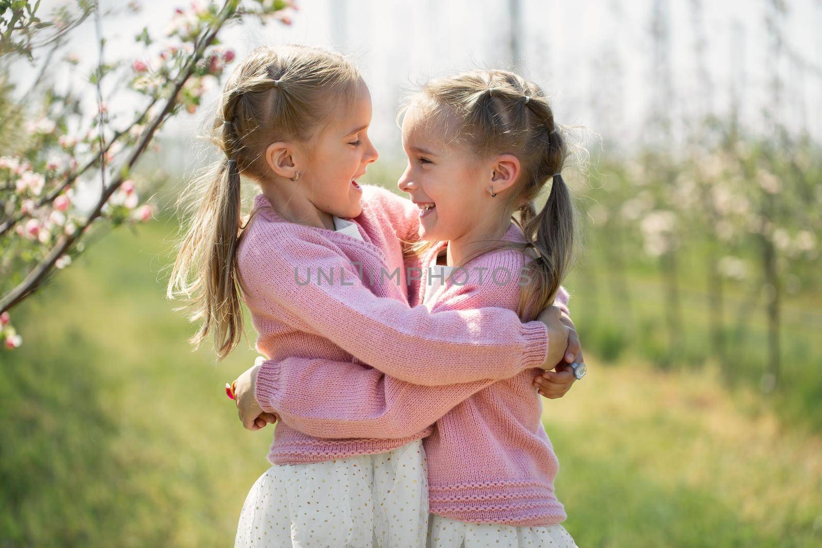 Happy twin sisters hug against the background of a green blooming Apple orchard. The sisters look at each other and laugh by StudioPeace