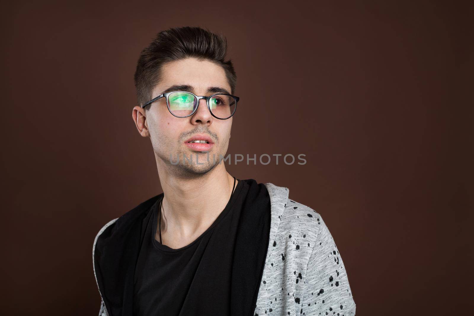 Young man in depression. Stylish male student wears round spectacles, has trendy hairstyle, looks confidently, isolated over brown background. People and human expressions