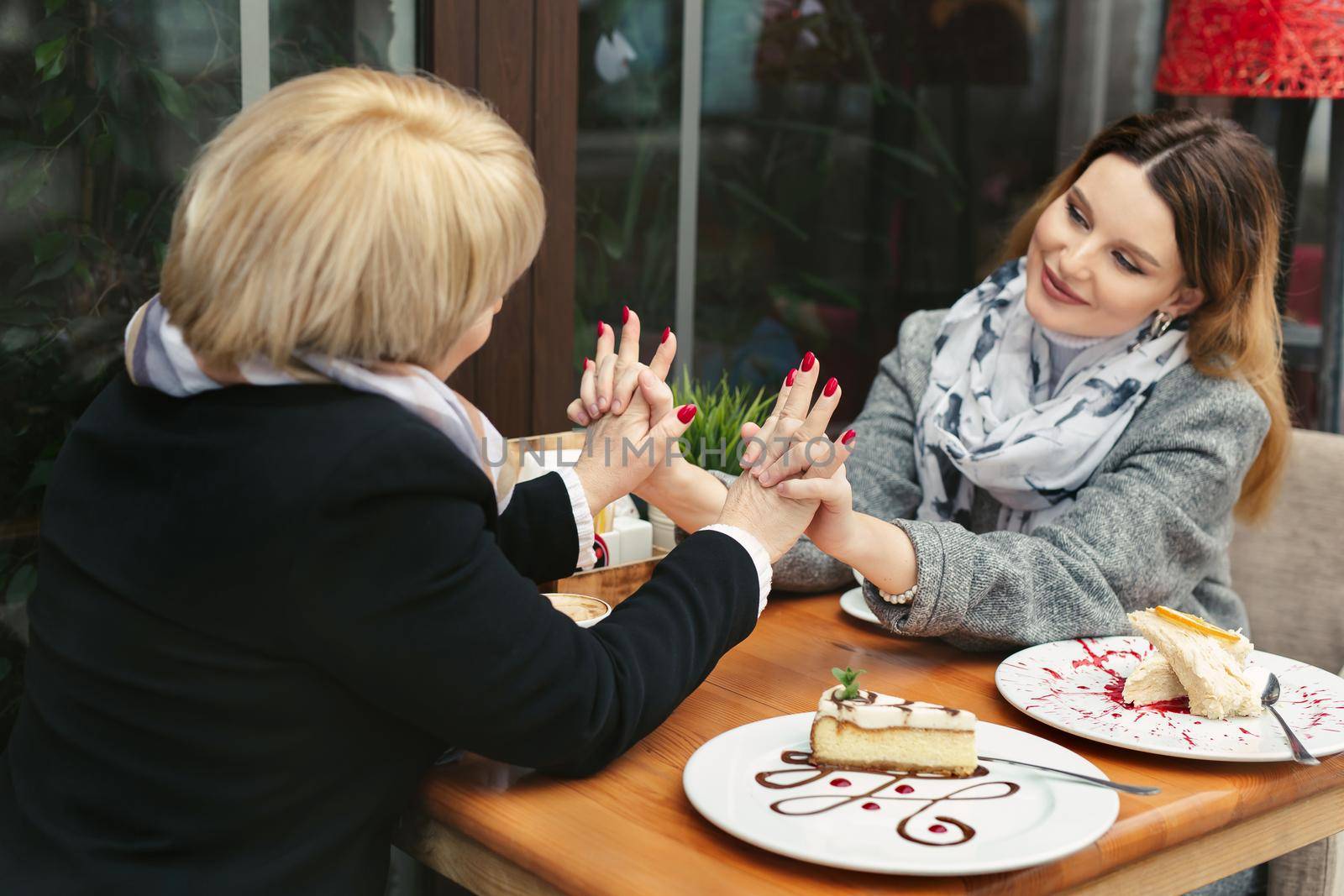 Family, an adult mother and daughter are sitting at a wooden table in a cafe on the street, looking at each other, smiling and holding hands by StudioPeace