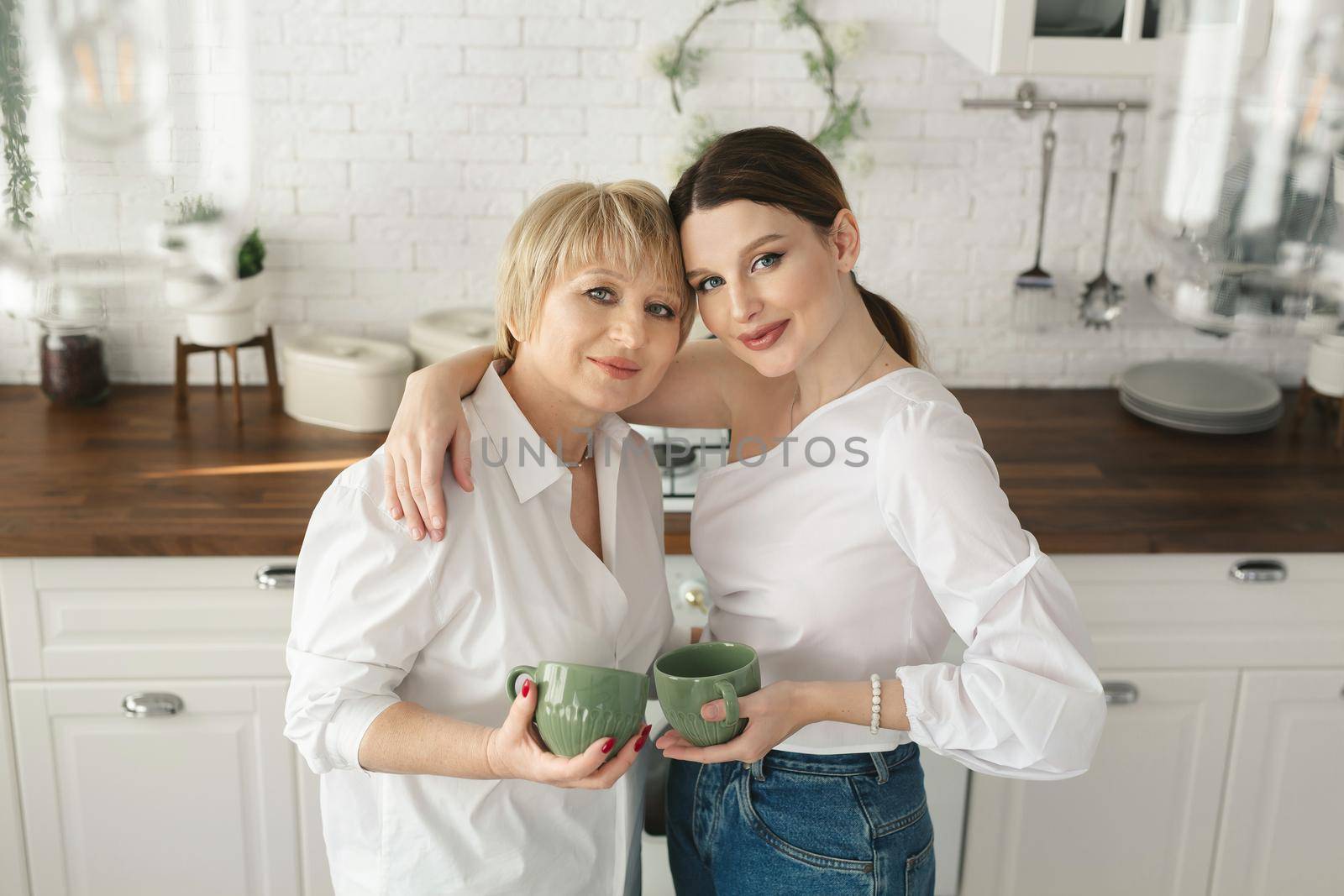 Portrait of a happy elderly mother and daughter in the kitchen, they drink tea and enjoy a conversation. Tender portrait of an old mother with an adult daughter at home