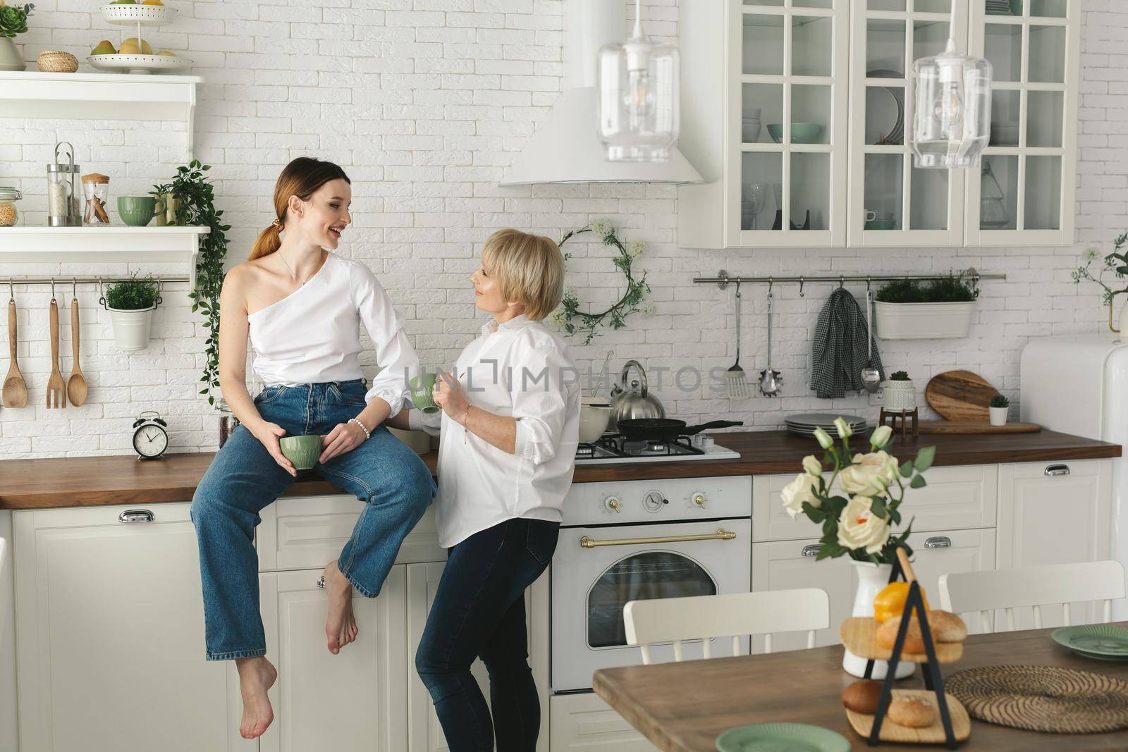 Happy elderly mother and daughter in the kitchen, they drink tea and enjoy a conversation. Old mother with an adult daughter at home. by StudioPeace