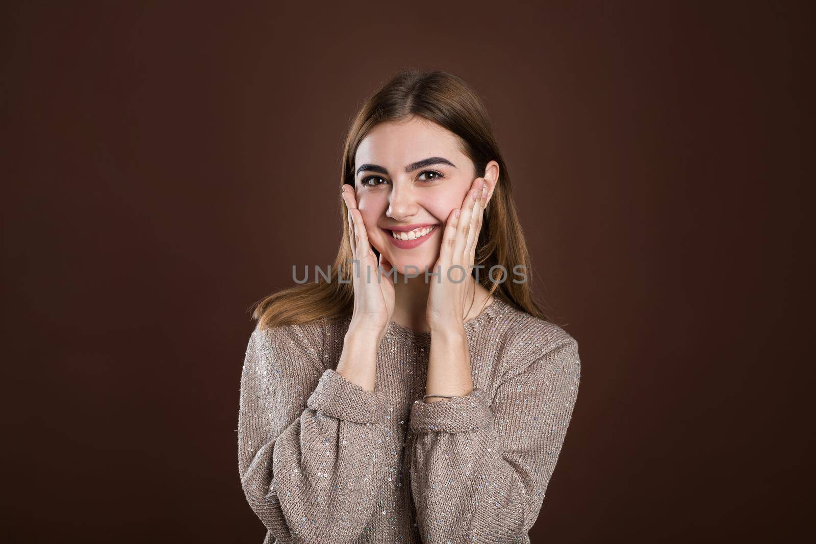Pleasantly surprised woman looking with bugged eyes holding hands on cheeks opening her mouth in amazement not expecting to recieve such great present. Happy model isolated over brown background. by StudioPeace