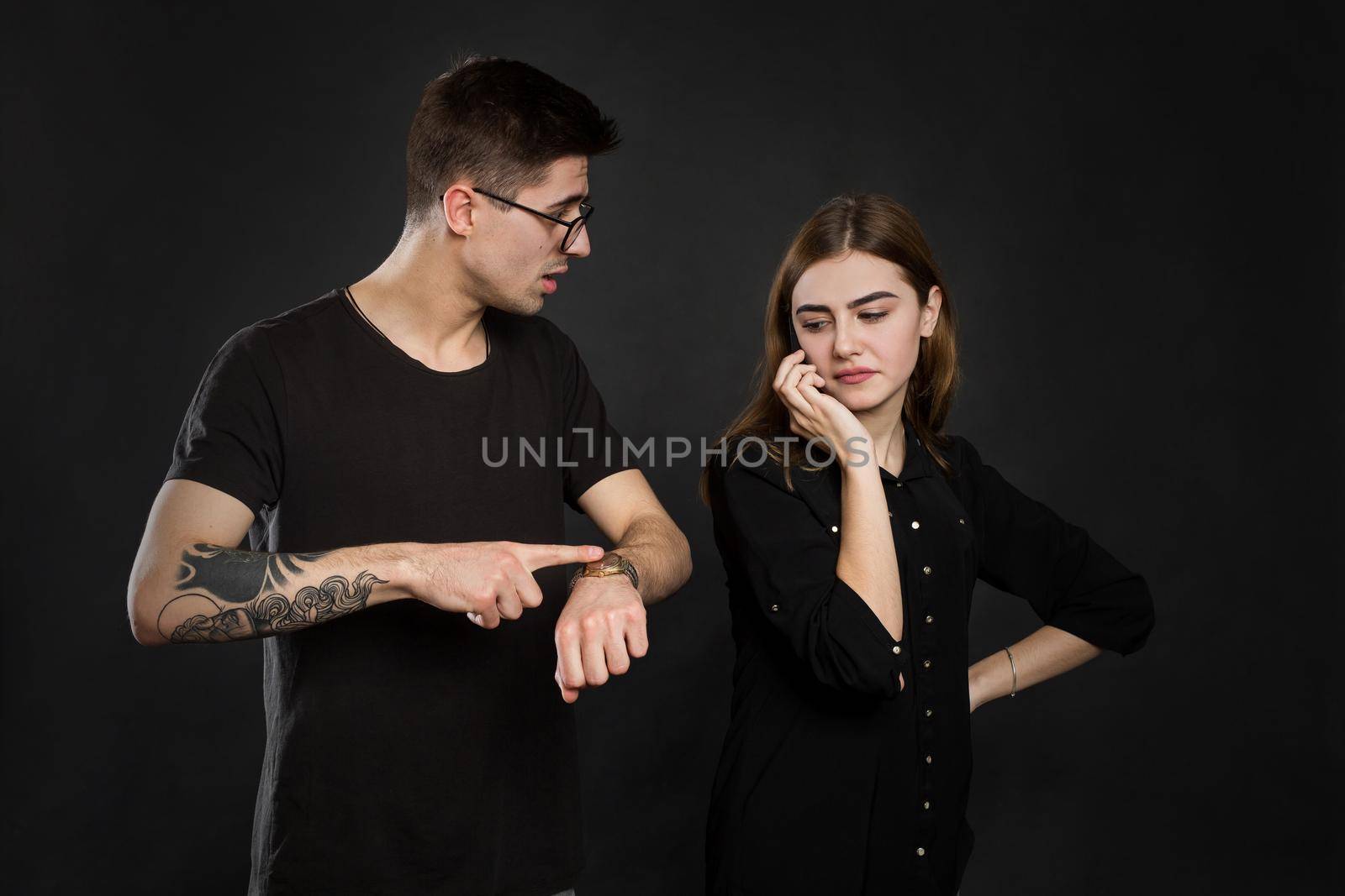 Portrait of a young couple standing with mobile phone, girl holding mobile phone while frustrated man standing near isolated over black background