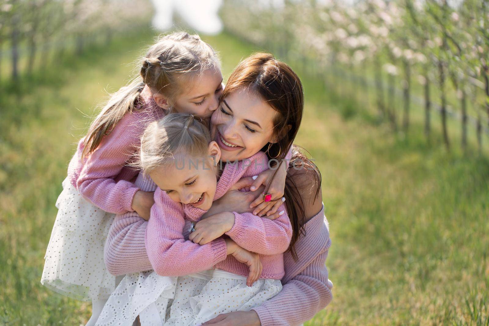 Portrait of a mother and her twin daughters in a blooming Apple orchard. Girls kiss their mother.
