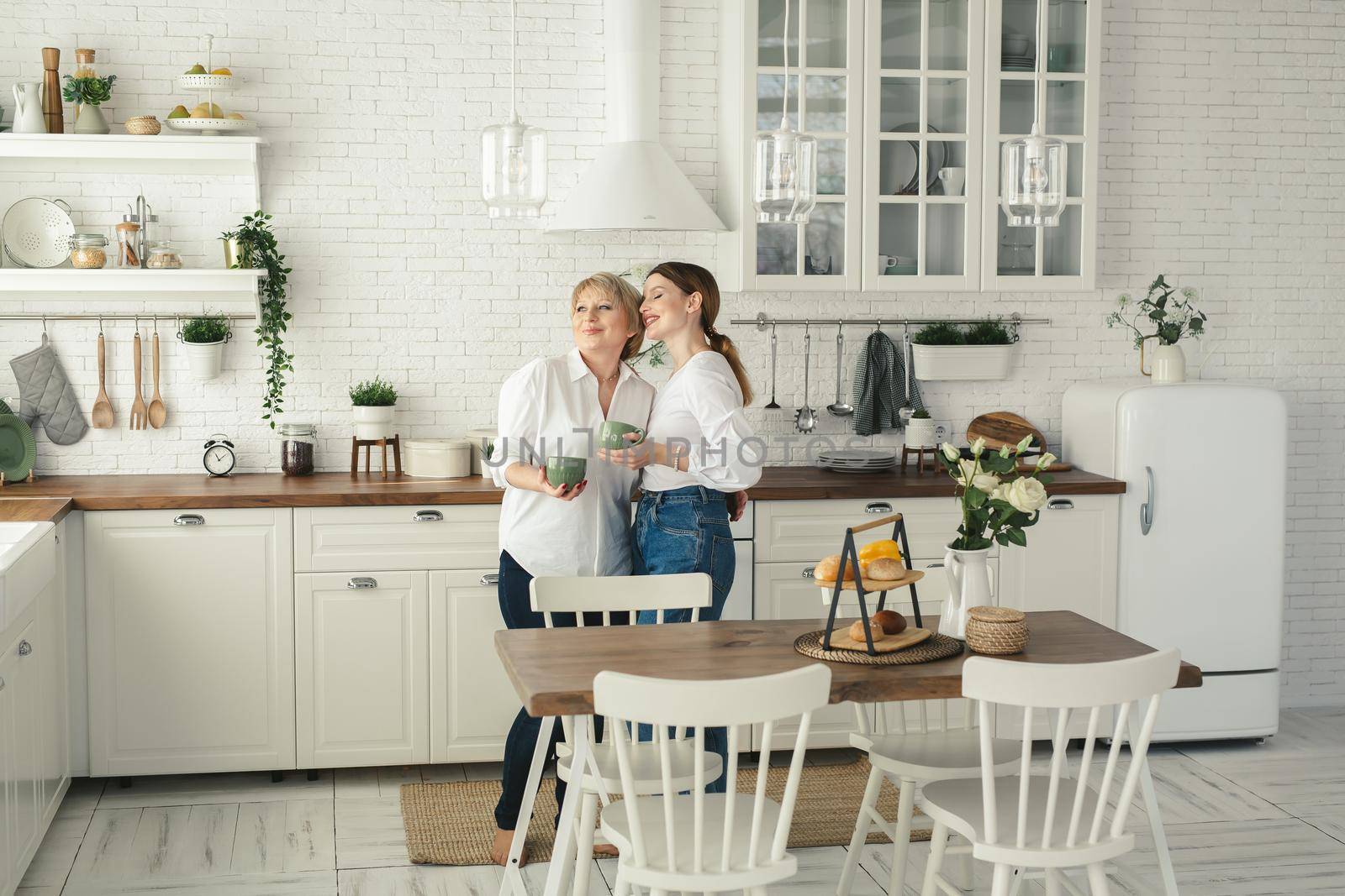 Portrait of a happy elderly mother and daughter in the kitchen, they drink tea and enjoy a conversation. Tender portrait of an old mother with an adult daughter at home. by StudioPeace