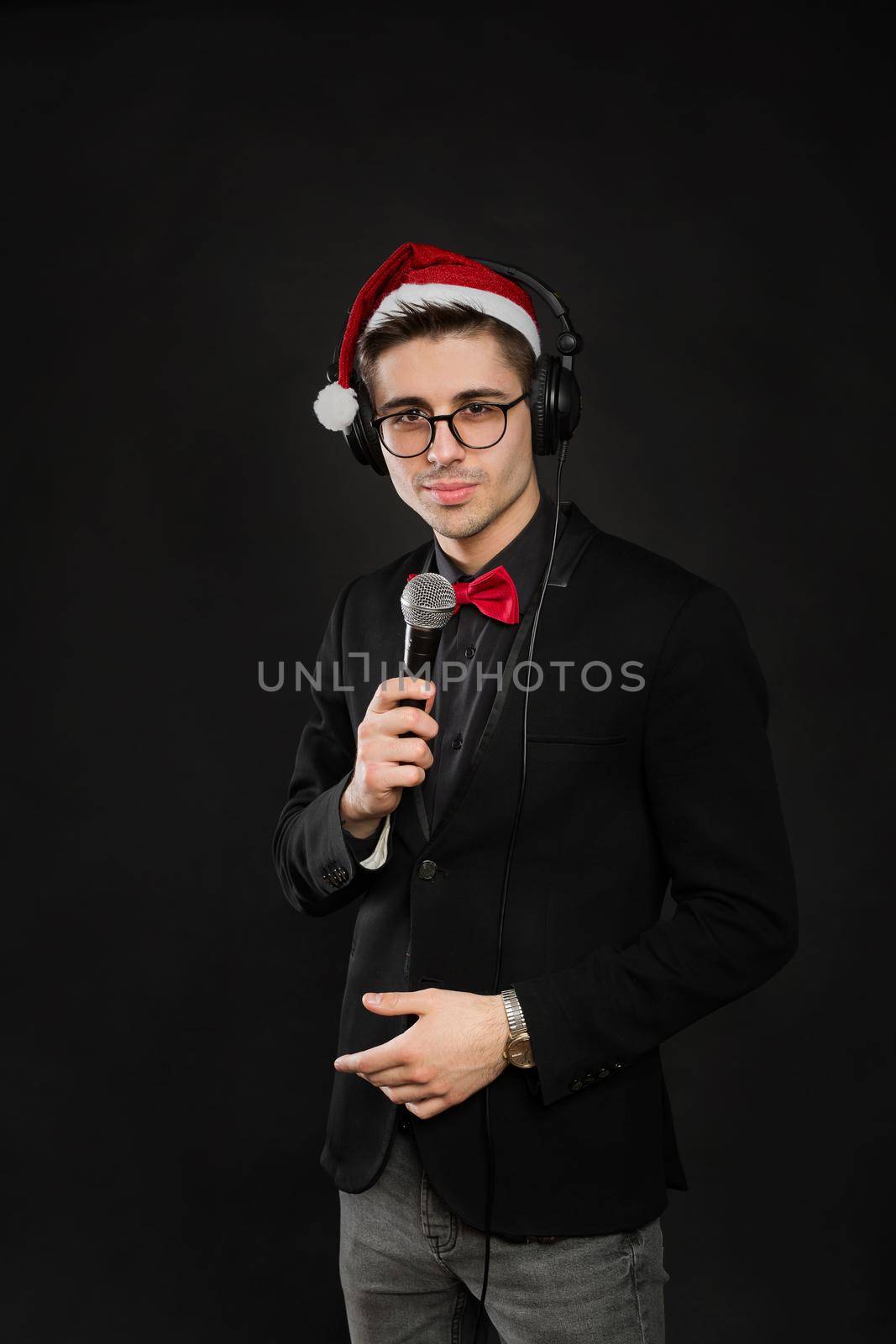 Portrait of a young male showman in glasses and a Santa Claus hat and headphones. With a microphone in his hands at a new year's party by StudioPeace