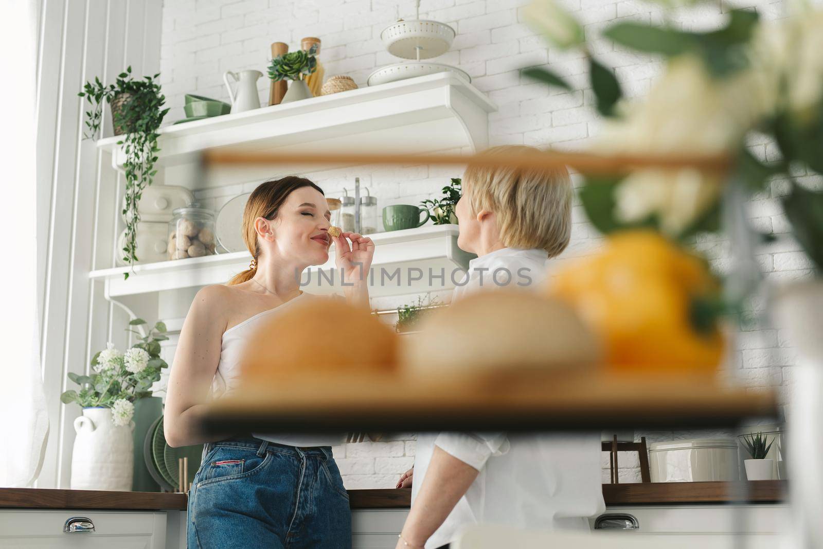 An adult daughter feeds her elderly mother fresh pastries in the kitchen by StudioPeace