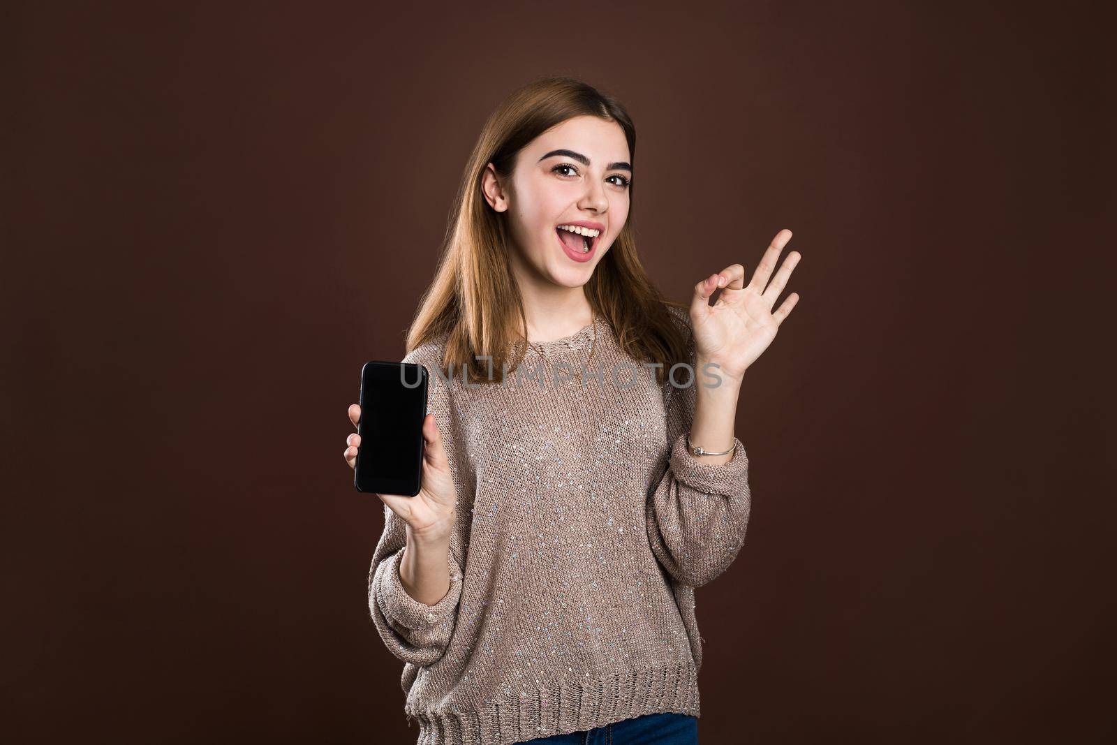 Smiling woman is pointing on smartphone standing on brown background. by StudioPeace