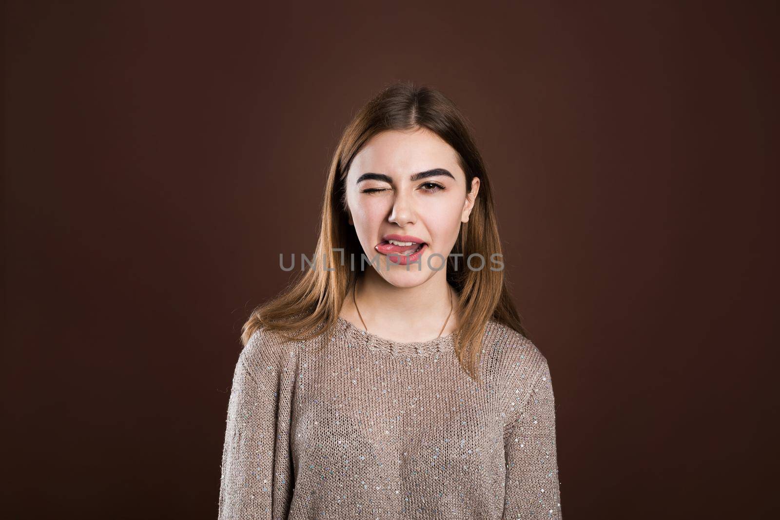 Cheerful young beautiful girl smiling showing tongue looking at camera over brown background. by StudioPeace