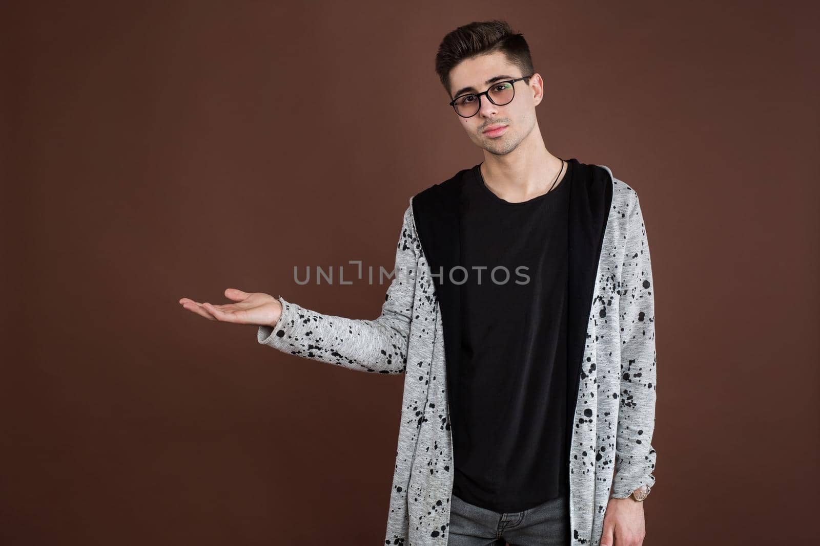 Handsome young adult man in shirt looking at camera and pointing away while standing isolated on brown background