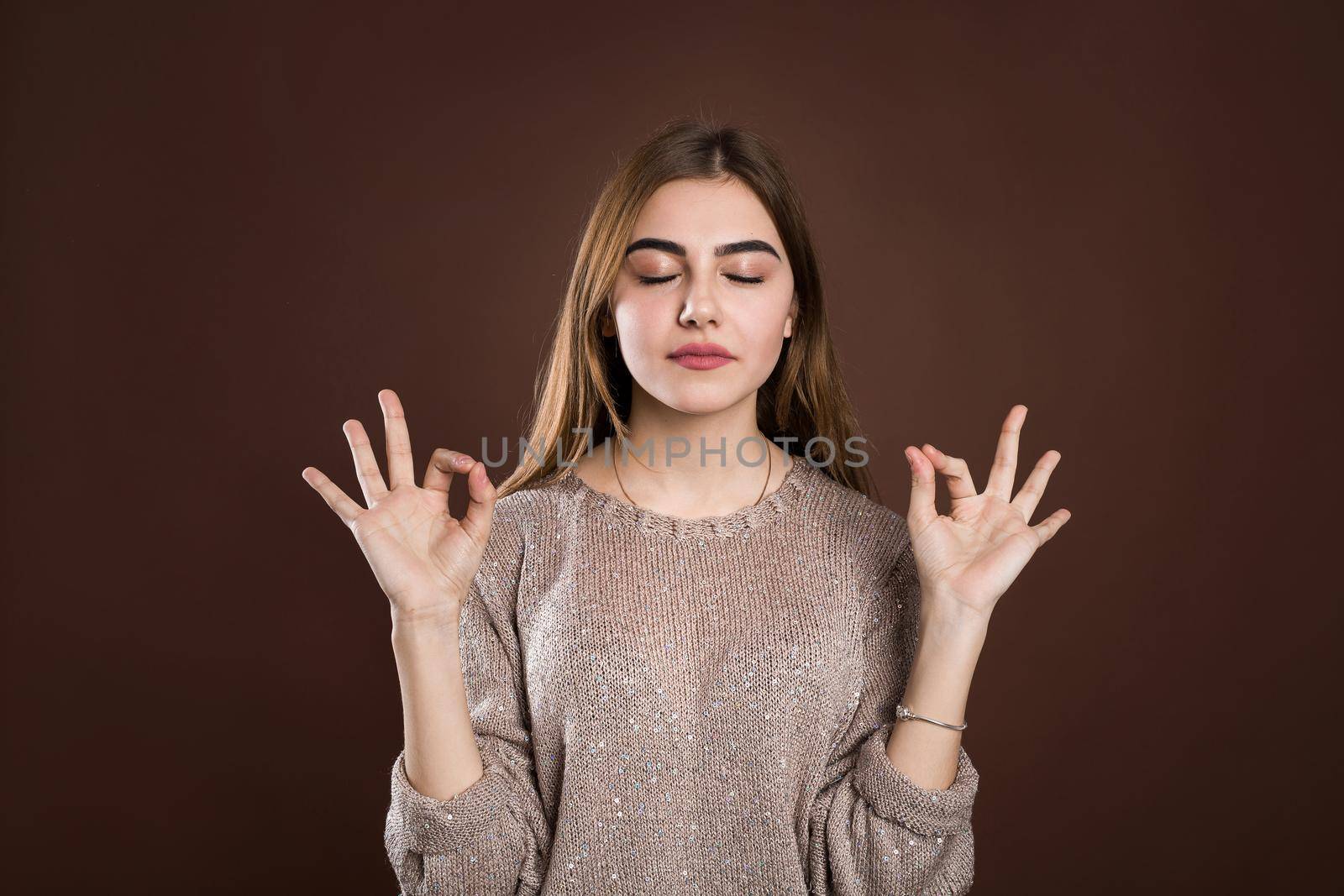 Portrait woman with calm and relaxed expression, standing in yoga pose with spreaded arms and zen signs over brown background. Girl feels relieved after morning meditation
