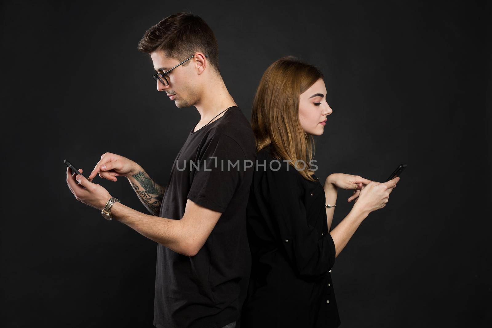 Profile portrait of young married couple, browsing information at their pdas, standing back to back, wearing casual outfits on black background. Another life in social nets. by StudioPeace