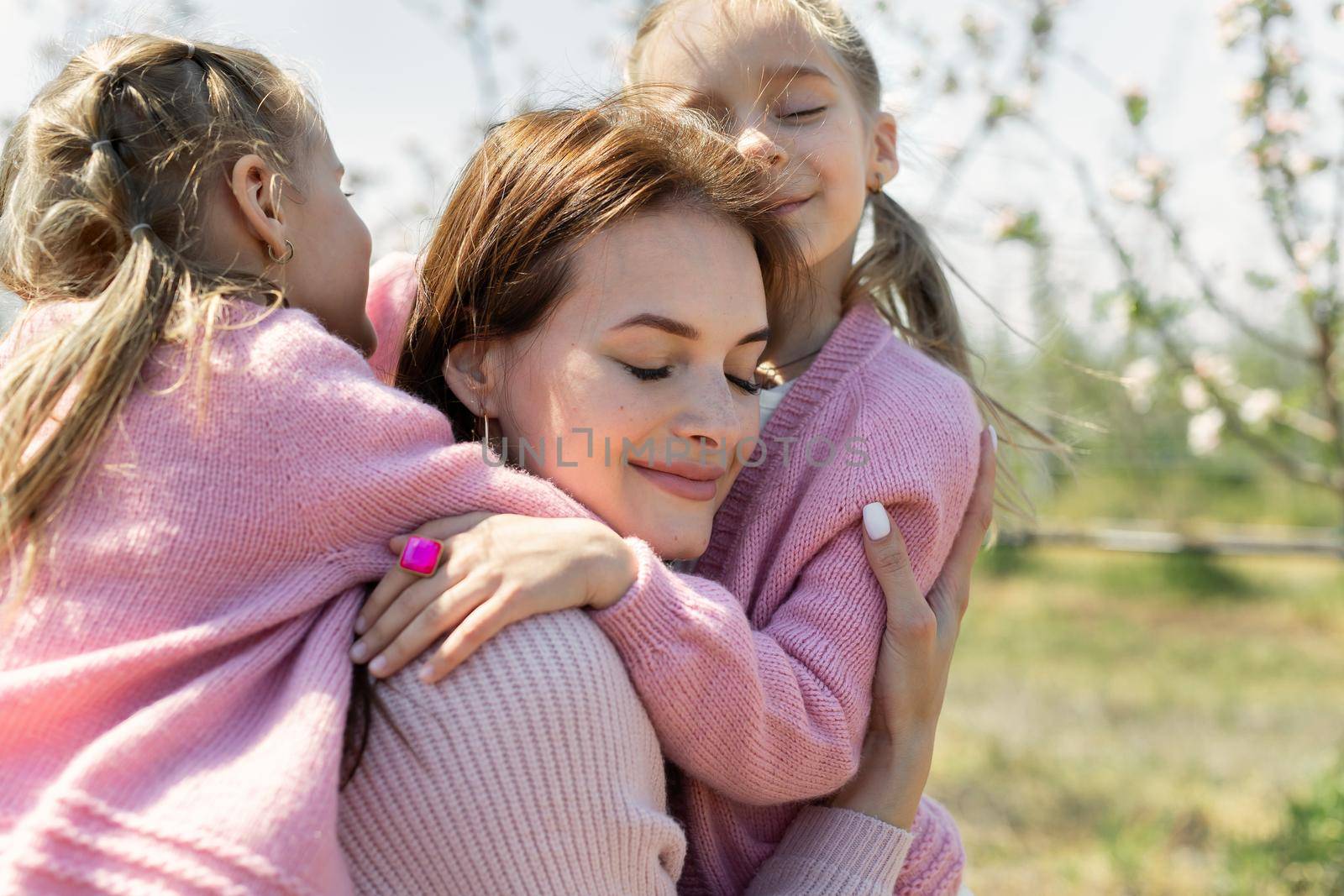 Close-up portrait of a young mother and her twin daughters hugging in an Apple orchard.