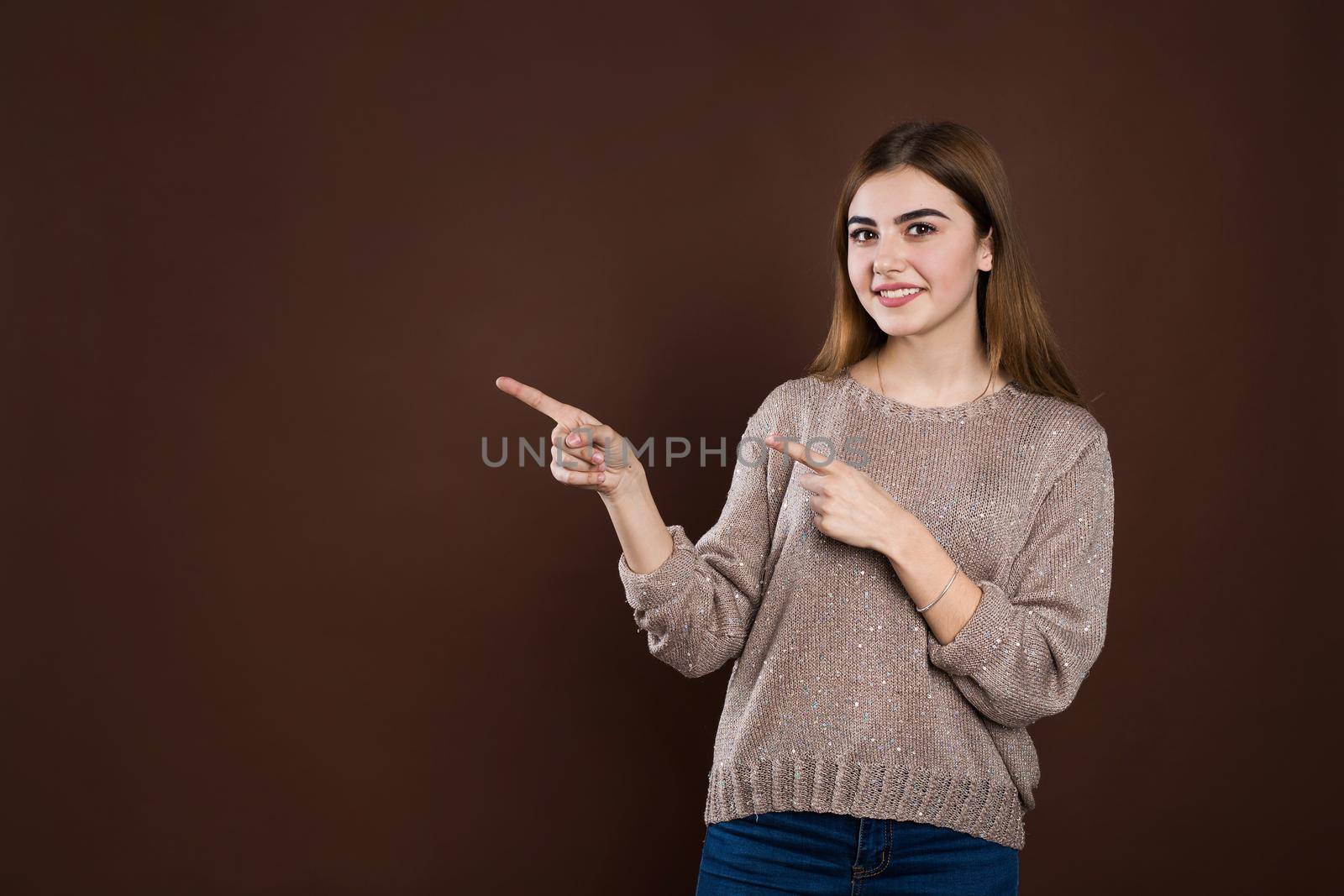 Smiling joyful woman in loose sweater posing against brown studio wall pointing at copy space for advertisment or promotional text. Positive emotions, feelings, joy, happiness. by StudioPeace