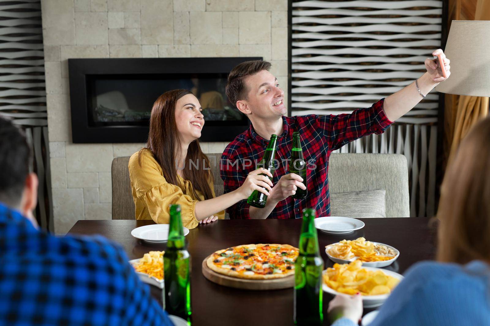 Group or company of young people - friends drink beer, eat pizza, talk and laugh and shoot selfie on the smartphone's camera on the background of the bar.