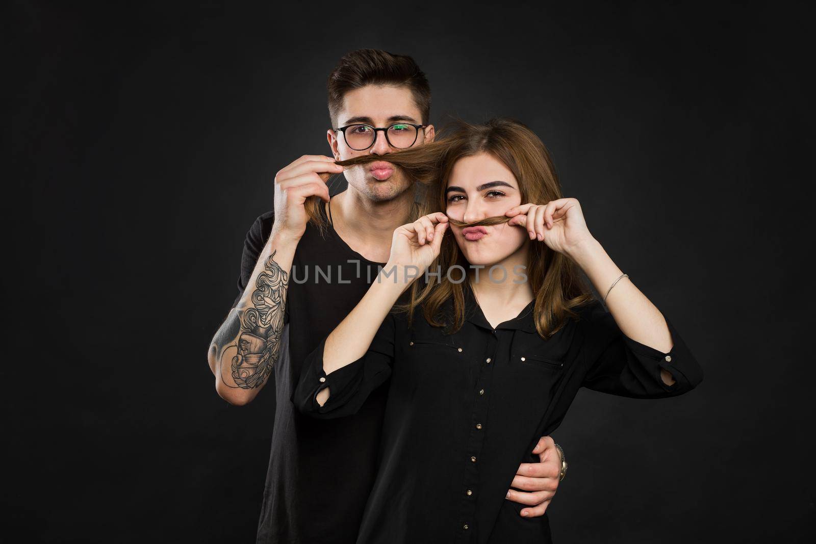 Beautiful young loving couple making fake moustache from hair while standing against black background. Funny moustache.