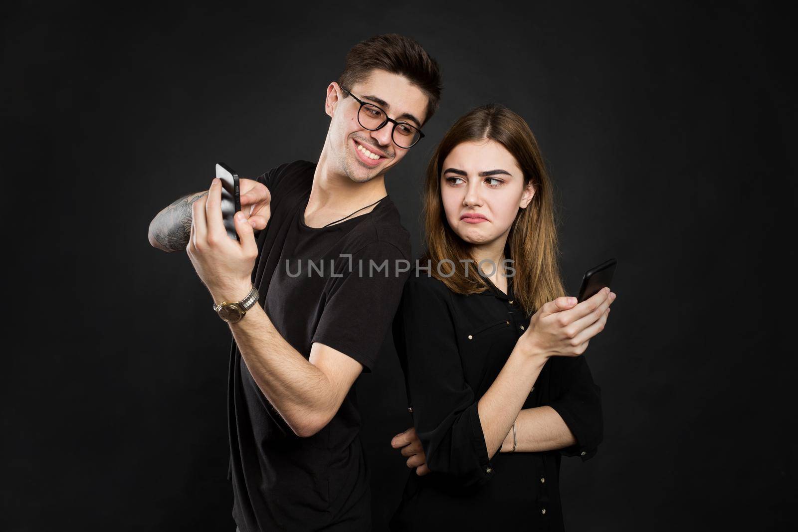 Portrait of a young couple standing with mobile phone, man is playing games on mobile phone while angry girl standing near and looking at camera isolated over black background