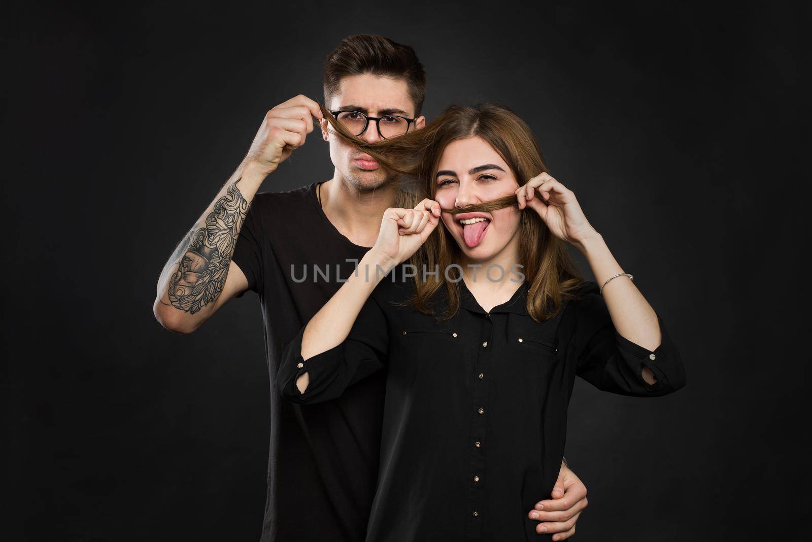 Beautiful young loving couple making fake moustache from hair while standing against black background. Funny moustache by StudioPeace