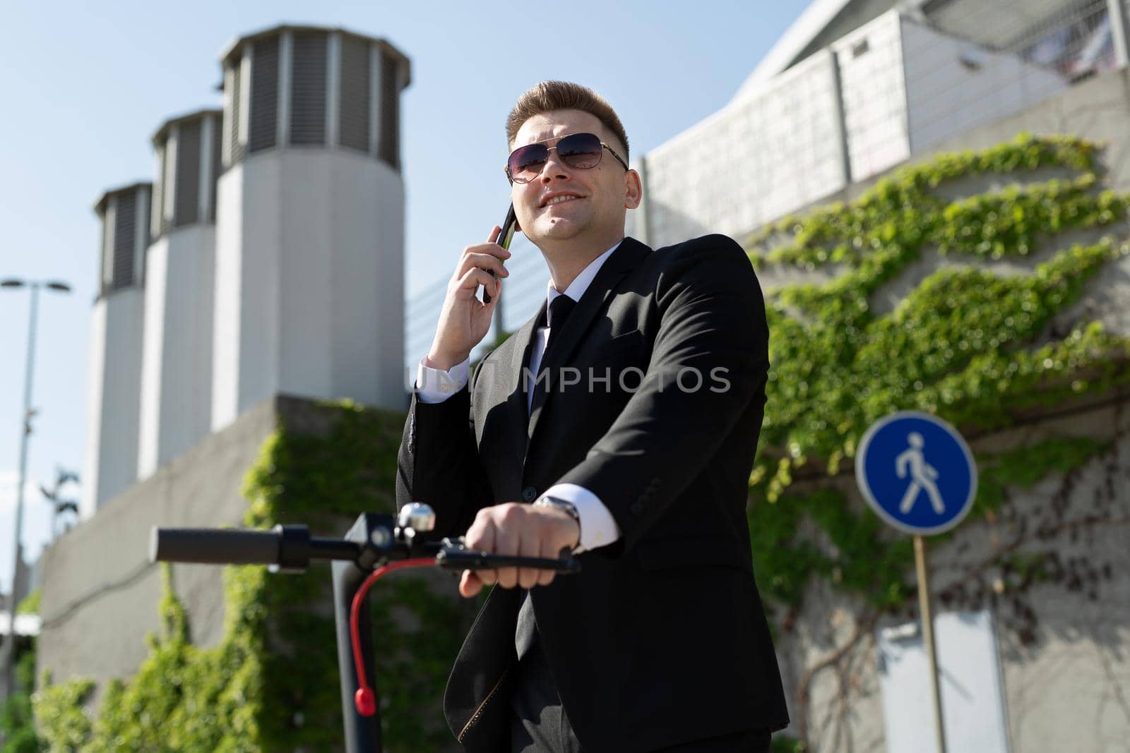 Man in a black business suit stands next to an electric scooter and talks on the phone.