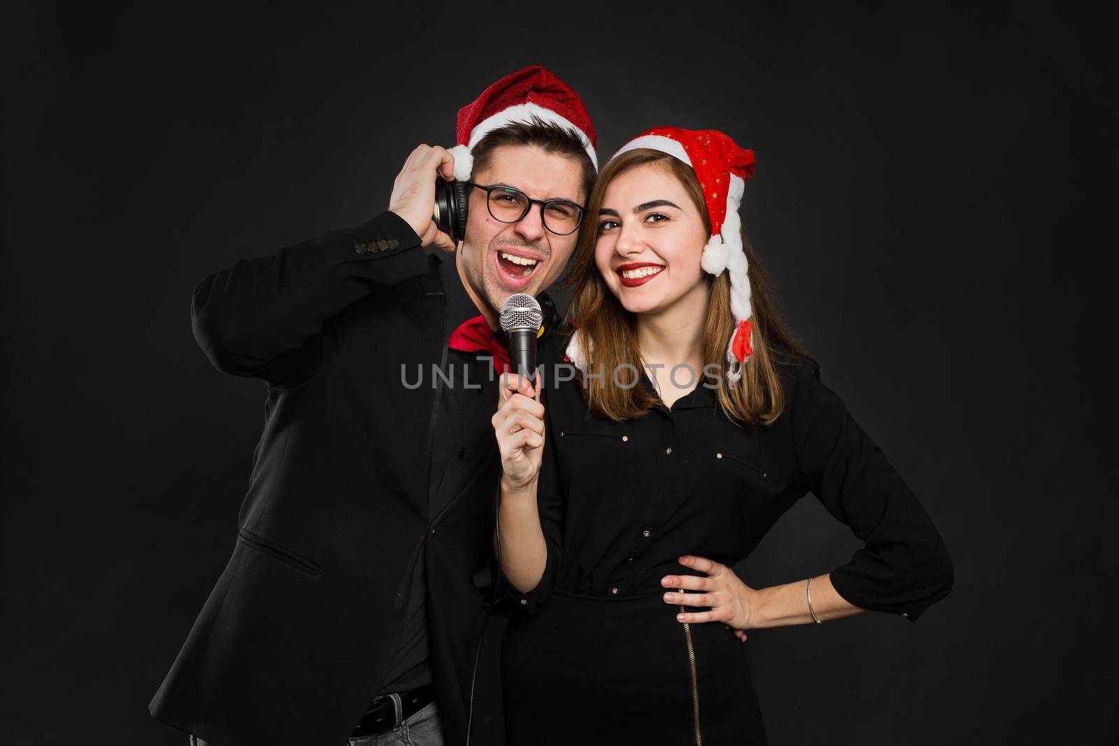 Young couple in love is enjoying a new year's party, putting on Santa hats and headphones, holding a microphone in their hands.