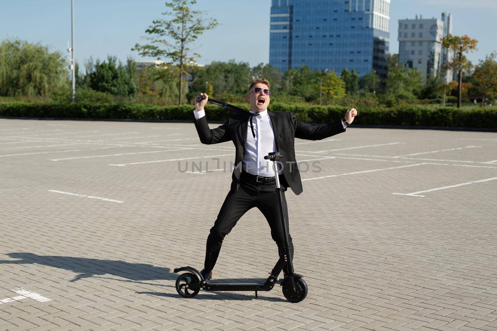 Young handsome businessman in a suit rides an electric scooter around the city and laughs by StudioPeace