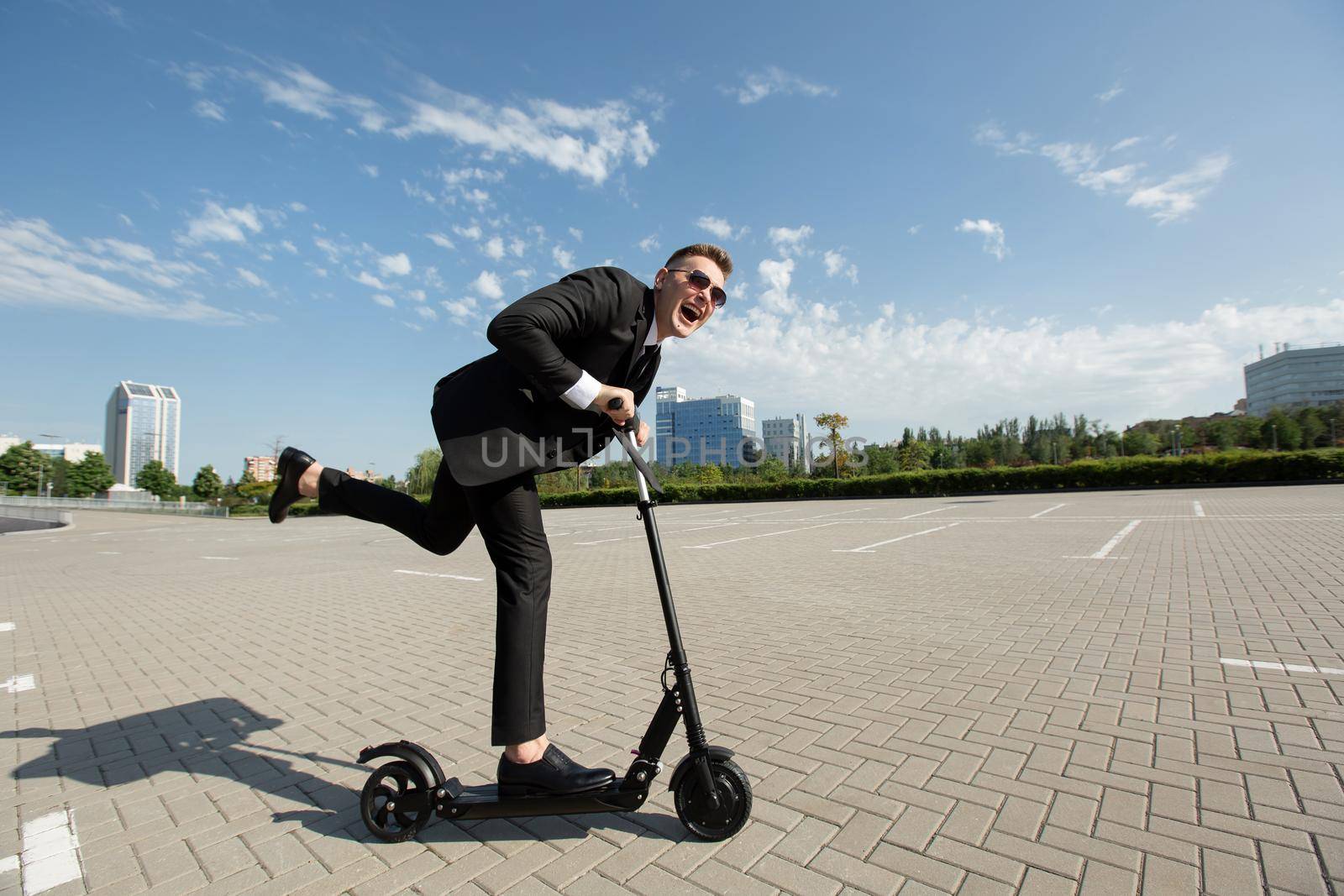Young handsome businessman in a suit rides an electric scooter around the city and laughs.