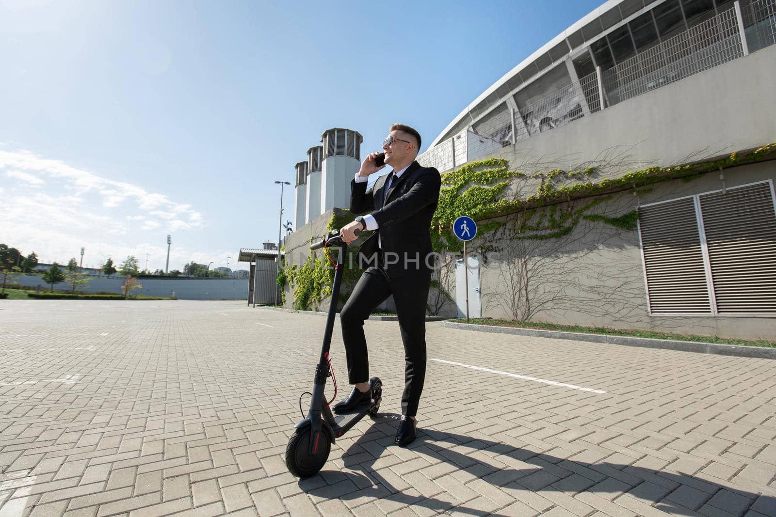 Man in a black business suit stands next to an electric scooter and talks on the phone by StudioPeace