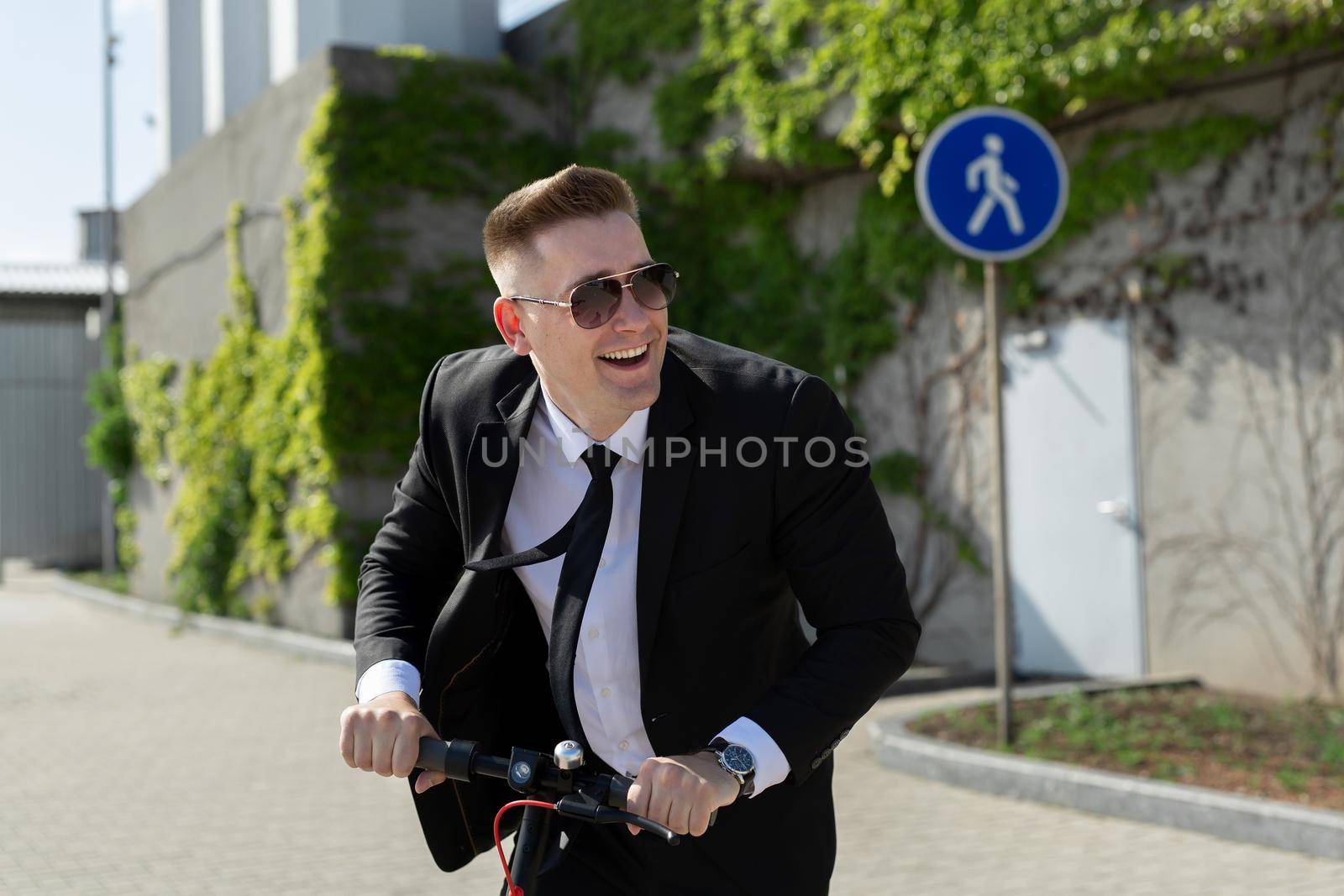 Man in a business suit and sunglasses rides an electric scooter and laughs.