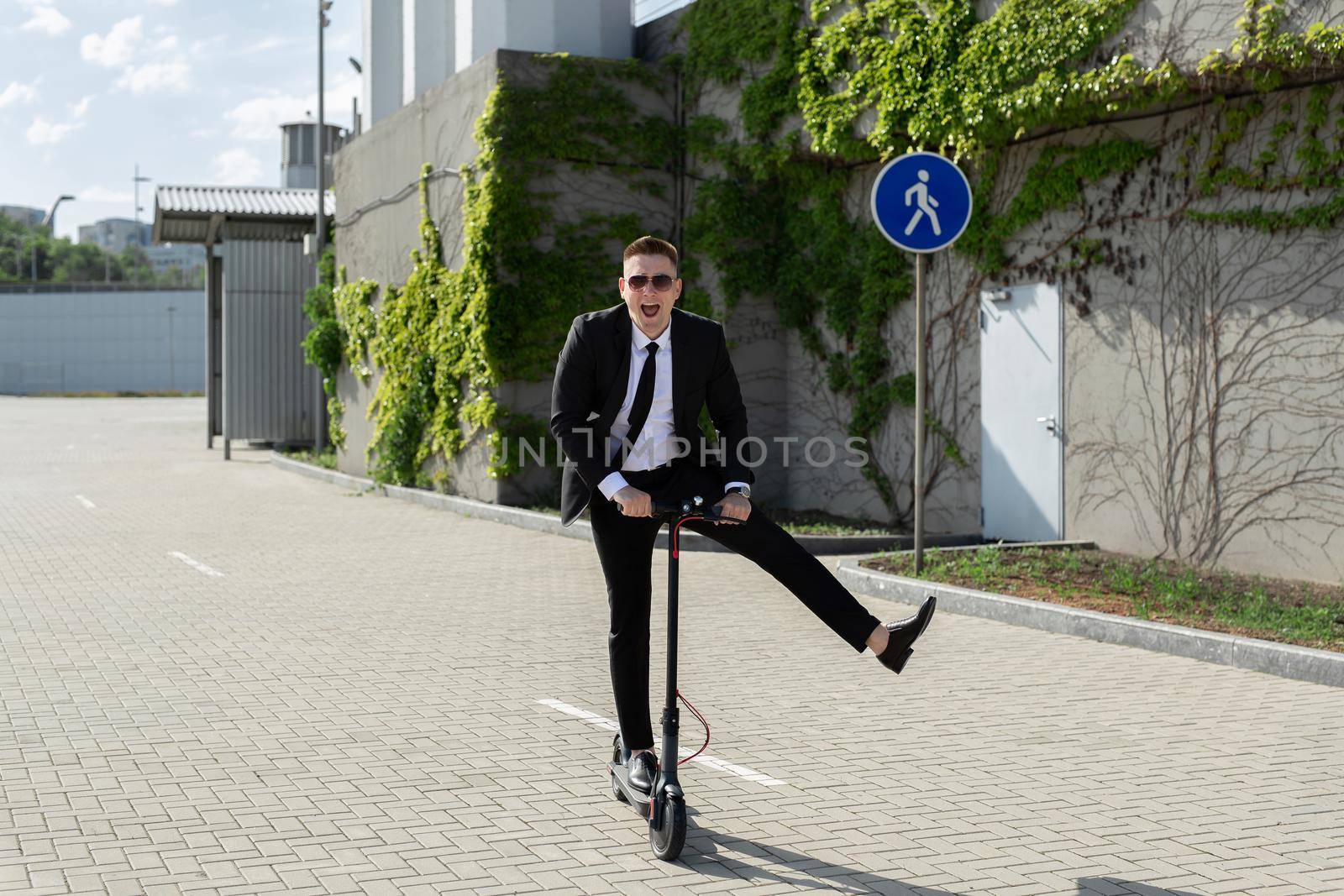 Young handsome businessman in a suit rides an electric scooter around the city and laughs.