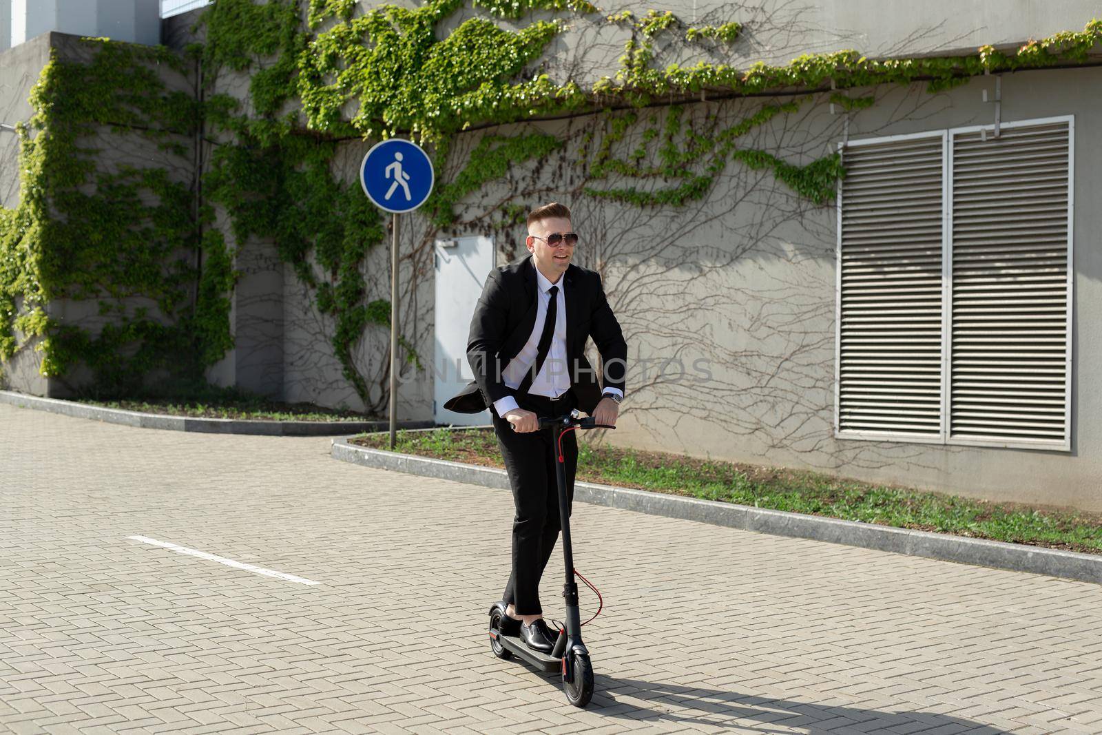 Young handsome businessman in a suit rides an electric scooter around the city and laughs.