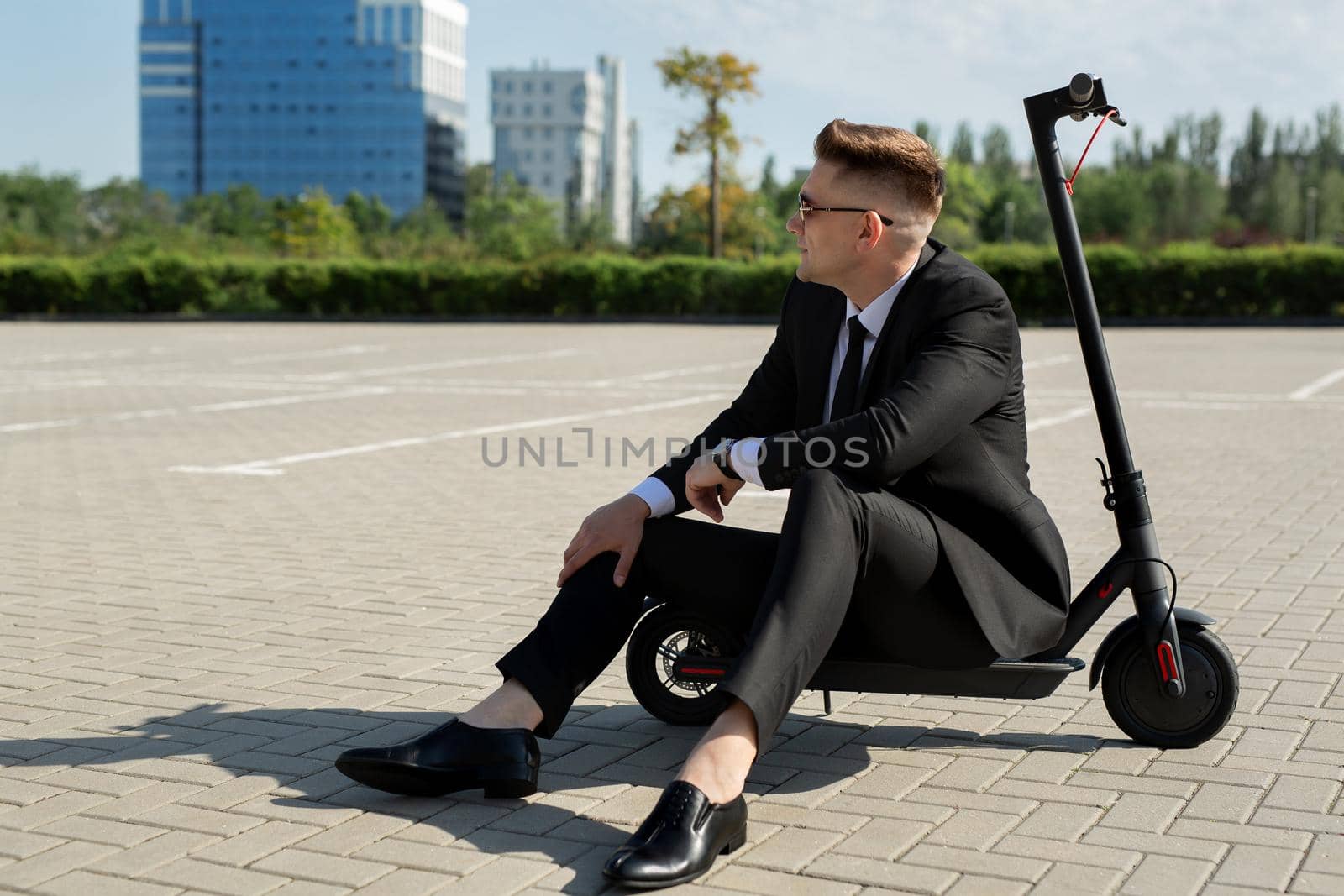 Young, handsome businessman in a suit sits on an electric scooter and laughs by StudioPeace