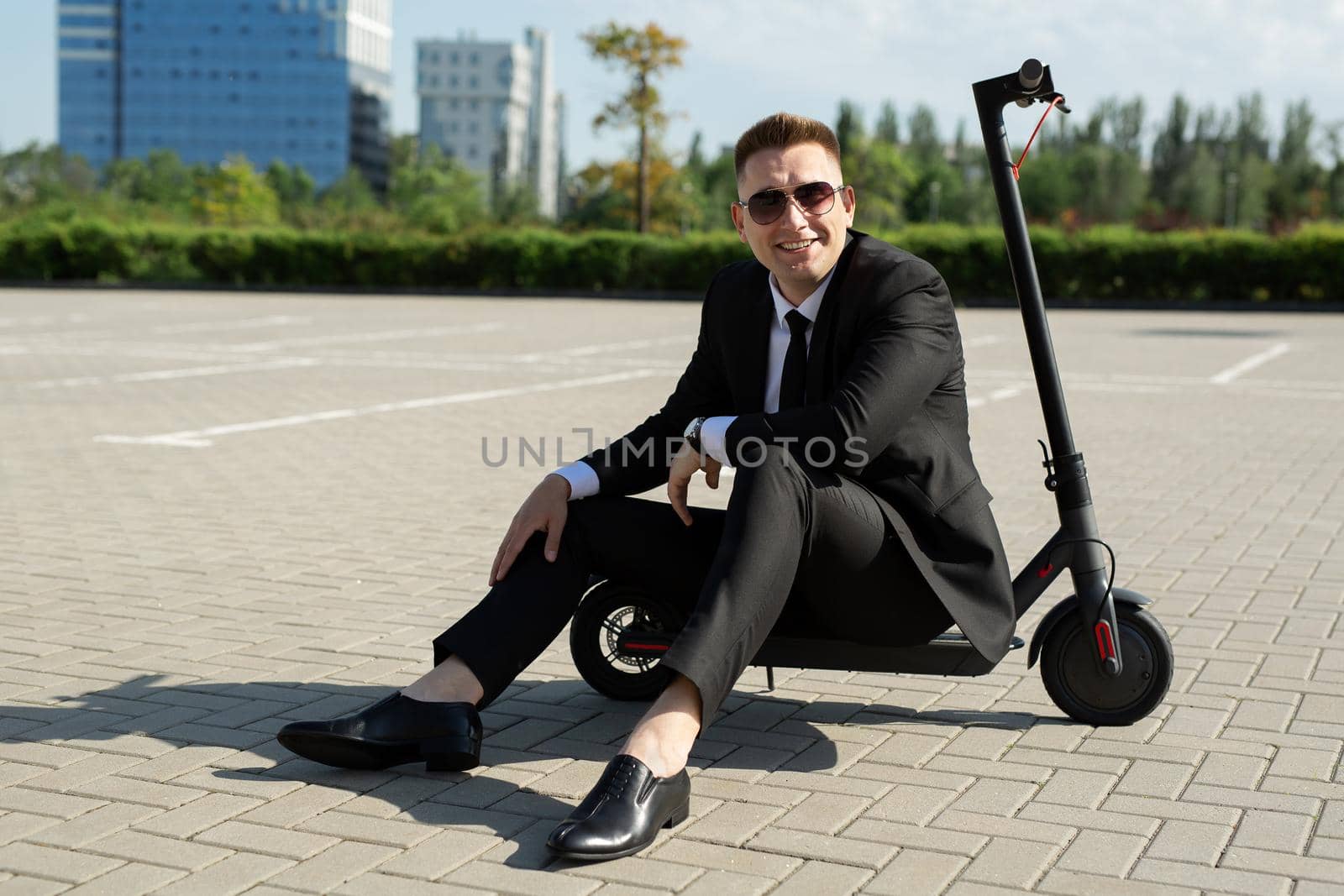 Young, handsome businessman in a suit sits on an electric scooter and laughs.