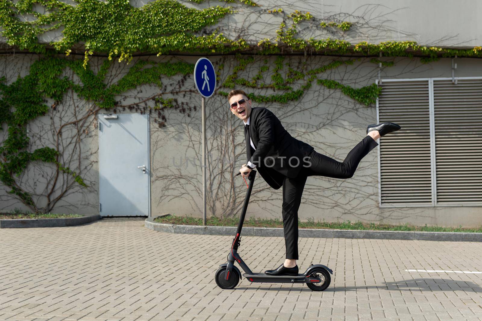 Man in a business suit and sunglasses rides an electric scooter on one and laughs by StudioPeace