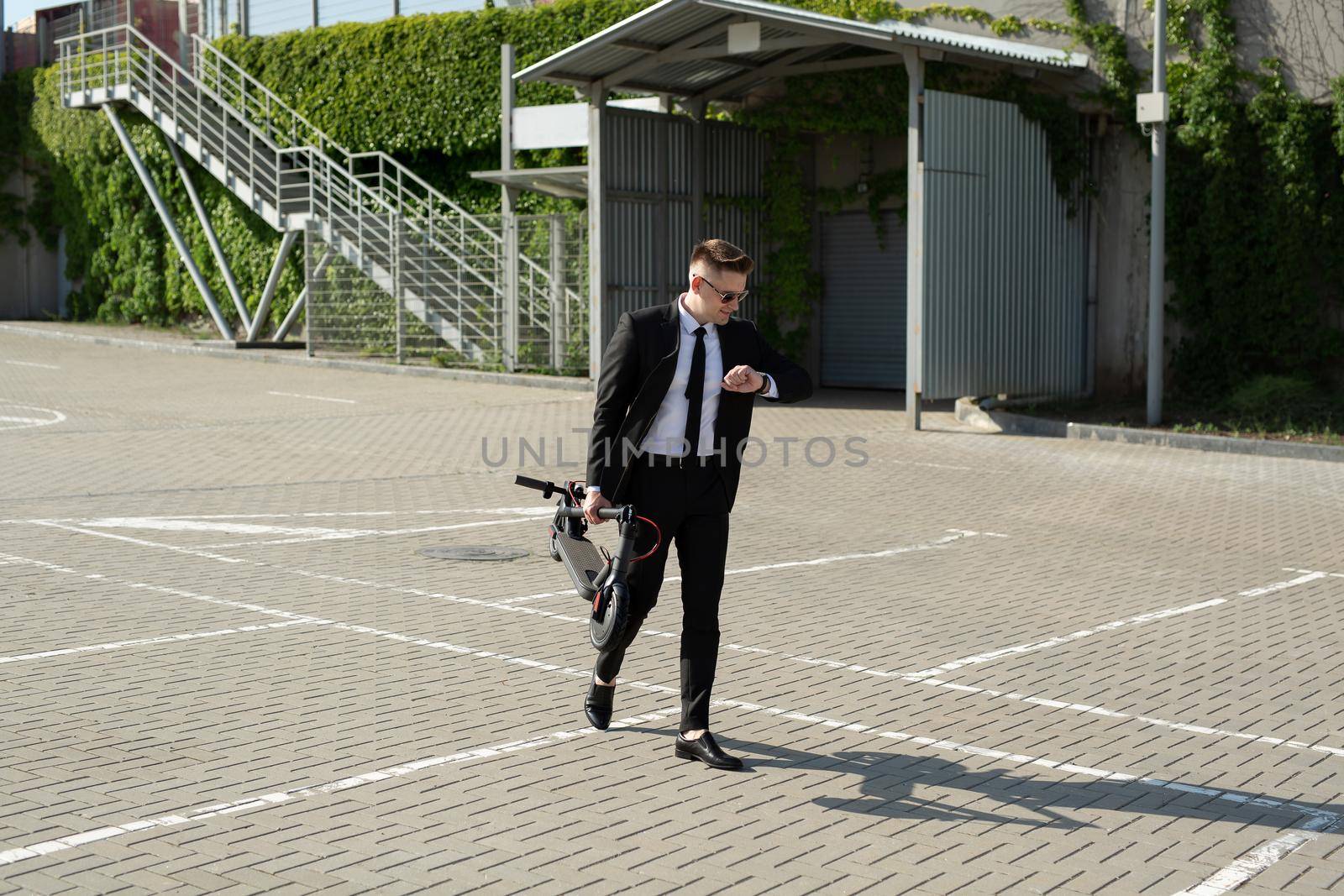 Young businessman in a suit looks at his watch and carries an electric scooter in his hands.