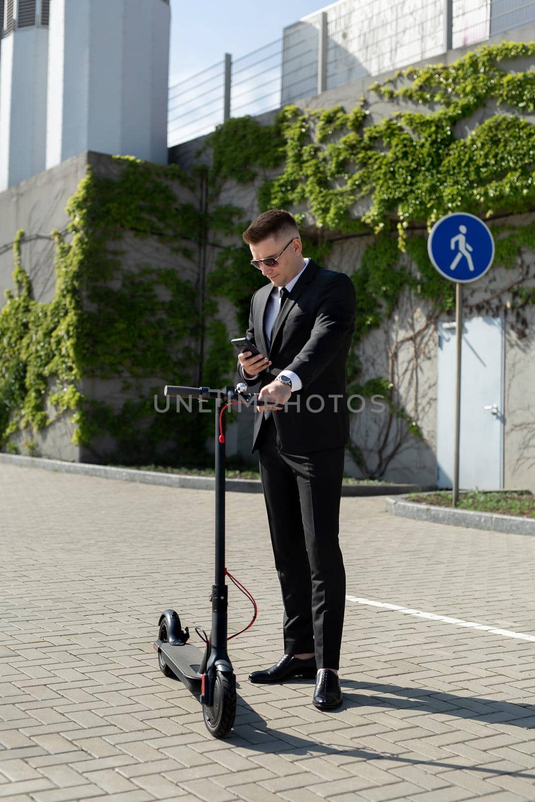 Man in a black business suit stands next to an electric scooter and holds a phone.