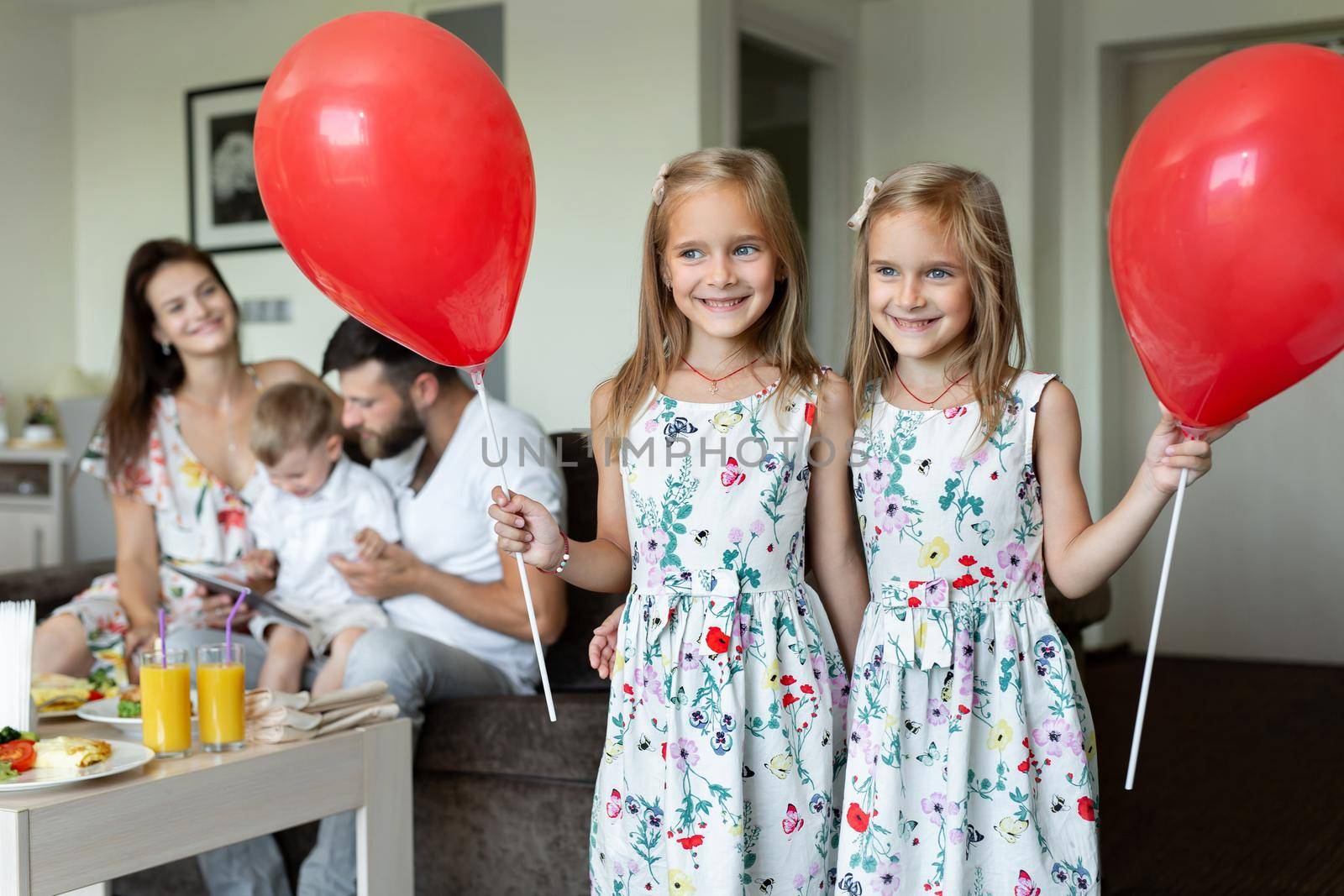 Two twin sisters pose with balloons in the background of the family. who eats breakfast in a hotel room by StudioPeace