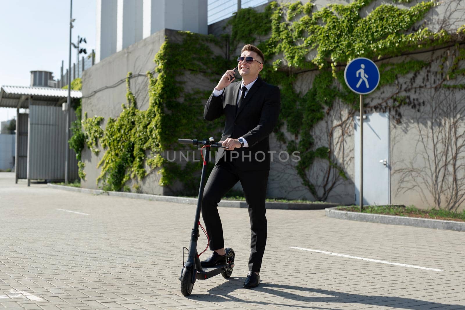 Man in a black business suit stands next to an electric scooter and talks on the phone.