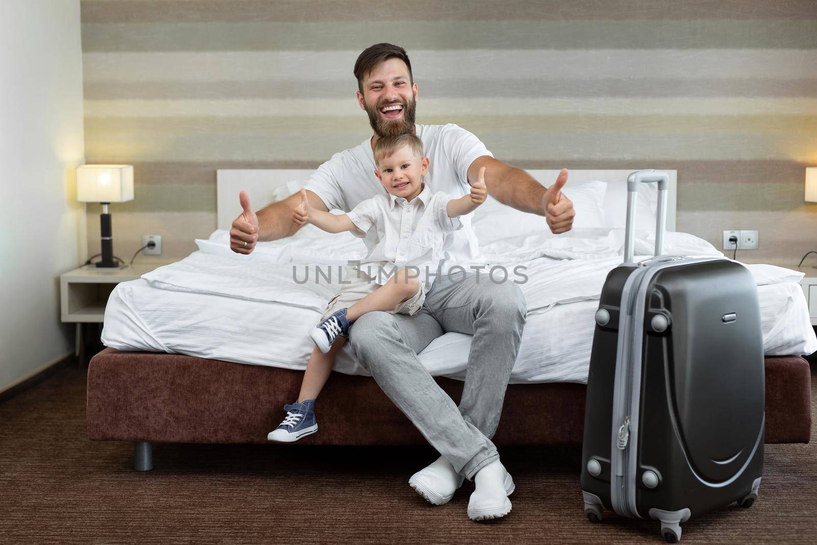 A young father and son sit on a bed in a hotel with a suitcase and laugh.