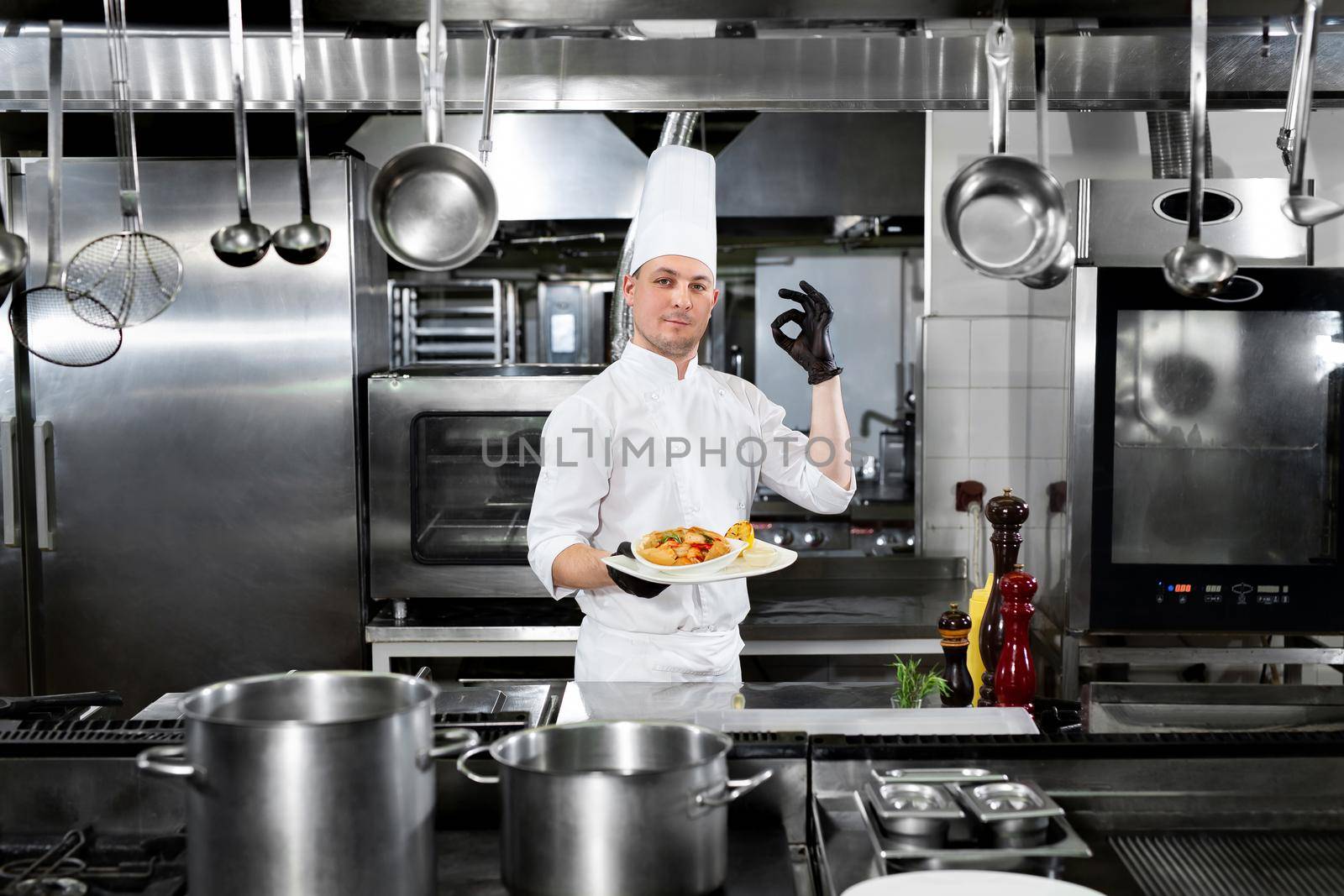 Chef in the restaurant holds a plate with a ready-made dish and shows the OK sign