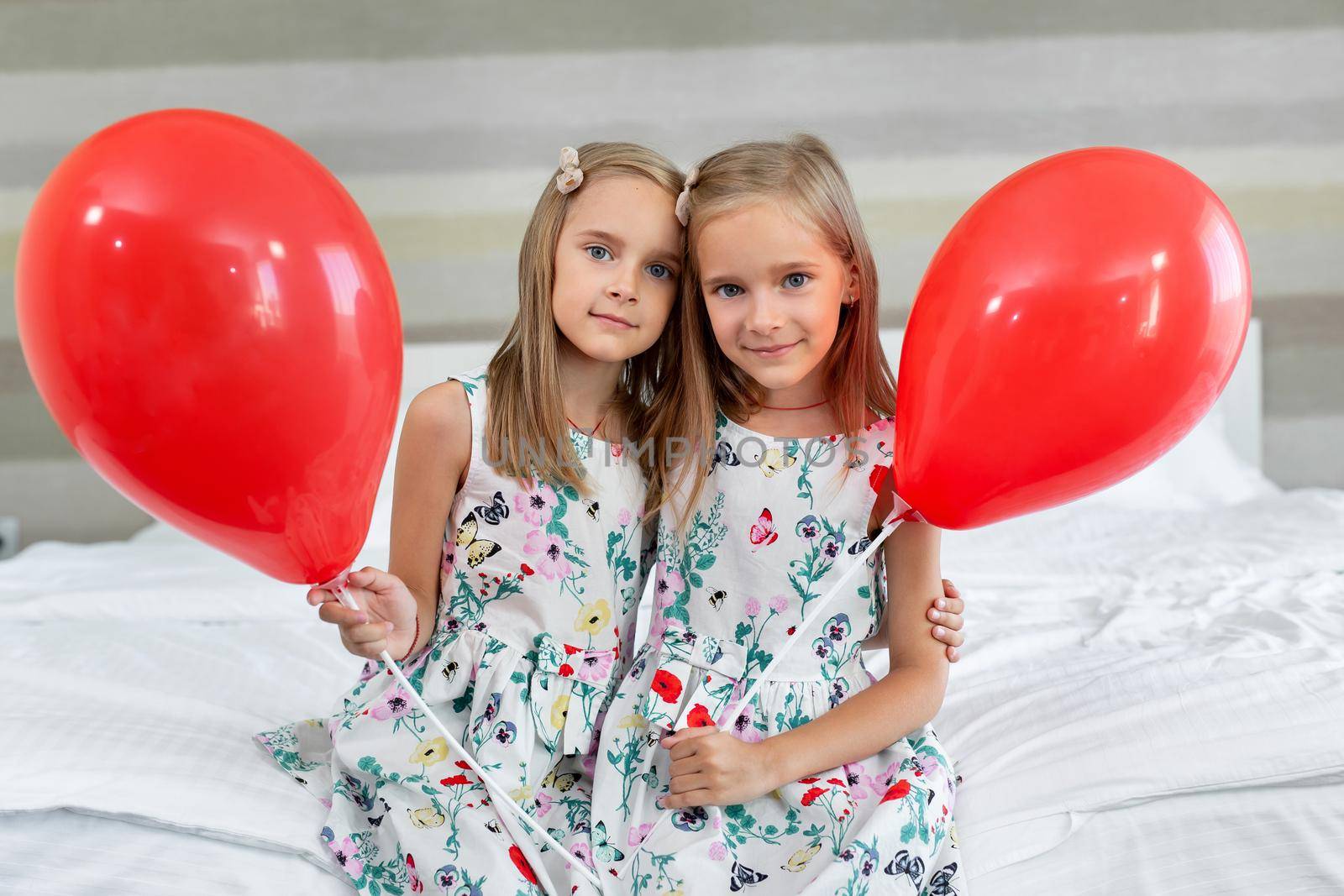 Two twin sisters in beautiful dresses are sitting on a bed in a hotel with balloons in their hands by StudioPeace