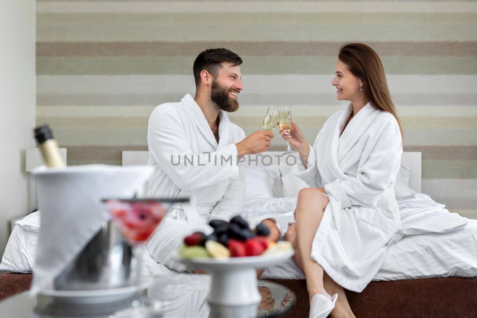 Happy couple a man and a woman in a white coat smile at each other and drink sparkling wine in bed in a hotel room by StudioPeace