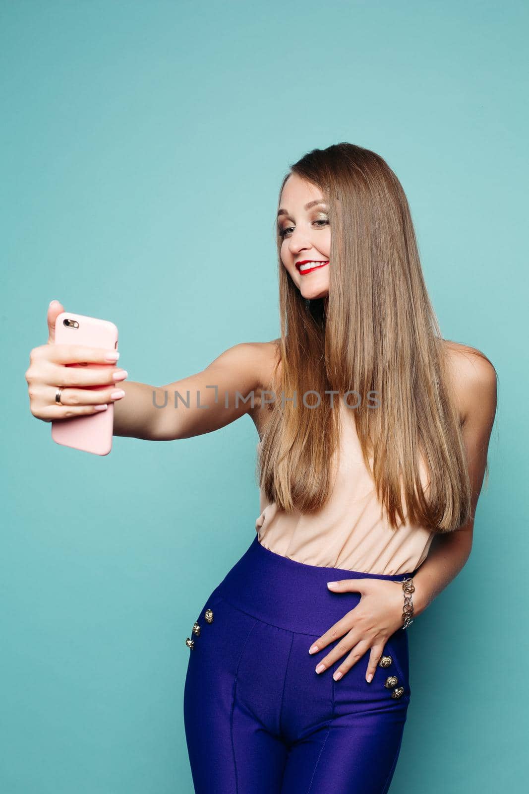 Studio portrait of beautiful elegant blonde woman with straight hair in blue trousers and top taking selfie with mobile phone over green background. Isolate.