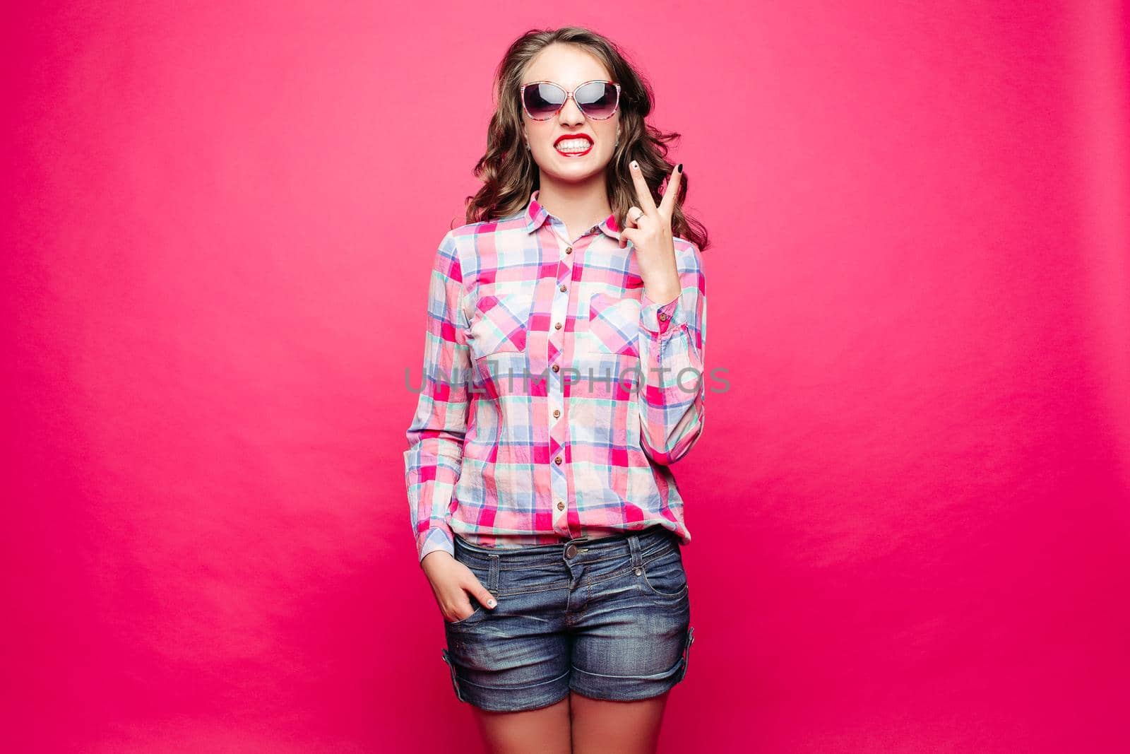 Studio portrait of funny girl in sunglasses showing peace sign with bare teeth over bright magenta background. Isolate.