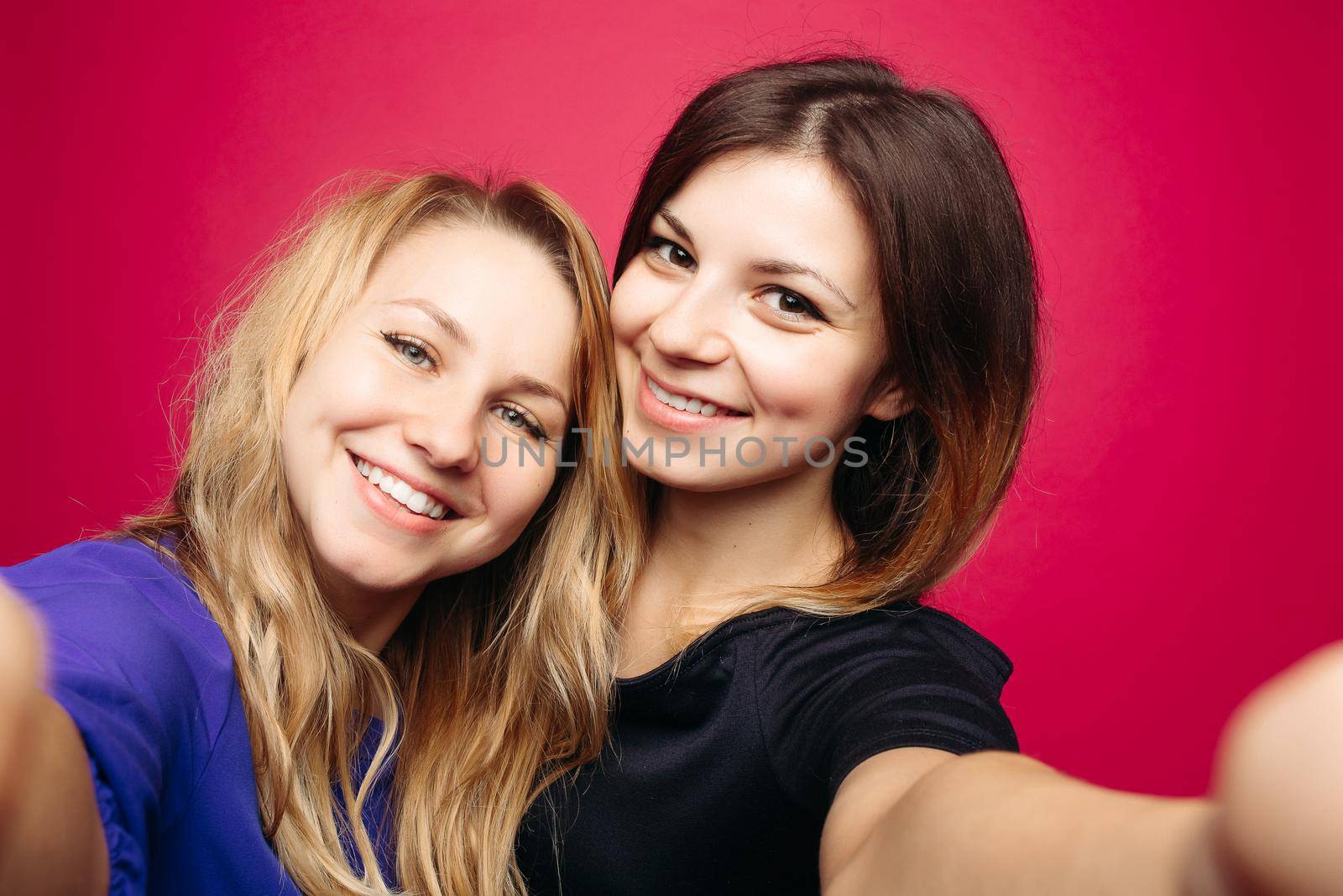 Self portrait of two beautiful, positivity girls, taking photo,smiling and posing together. Happy emotionally women with hairstyles,makeup, using camera at studio. Woman s friendship. Pink background.