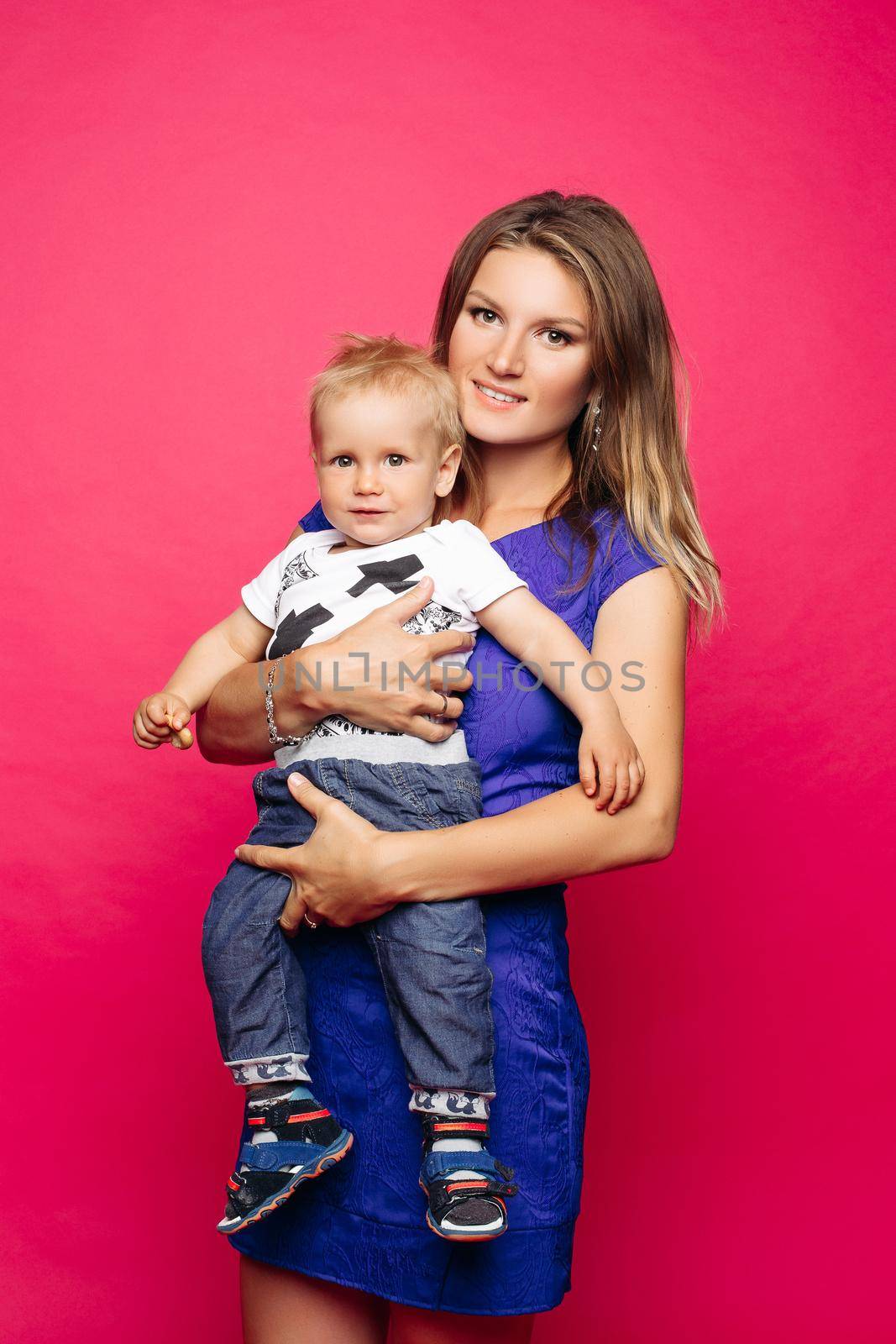 Studio portrait of loving mother with blonde hair embracing her playful baby over magenta background. Smiling at camera while baby moving in her hands.