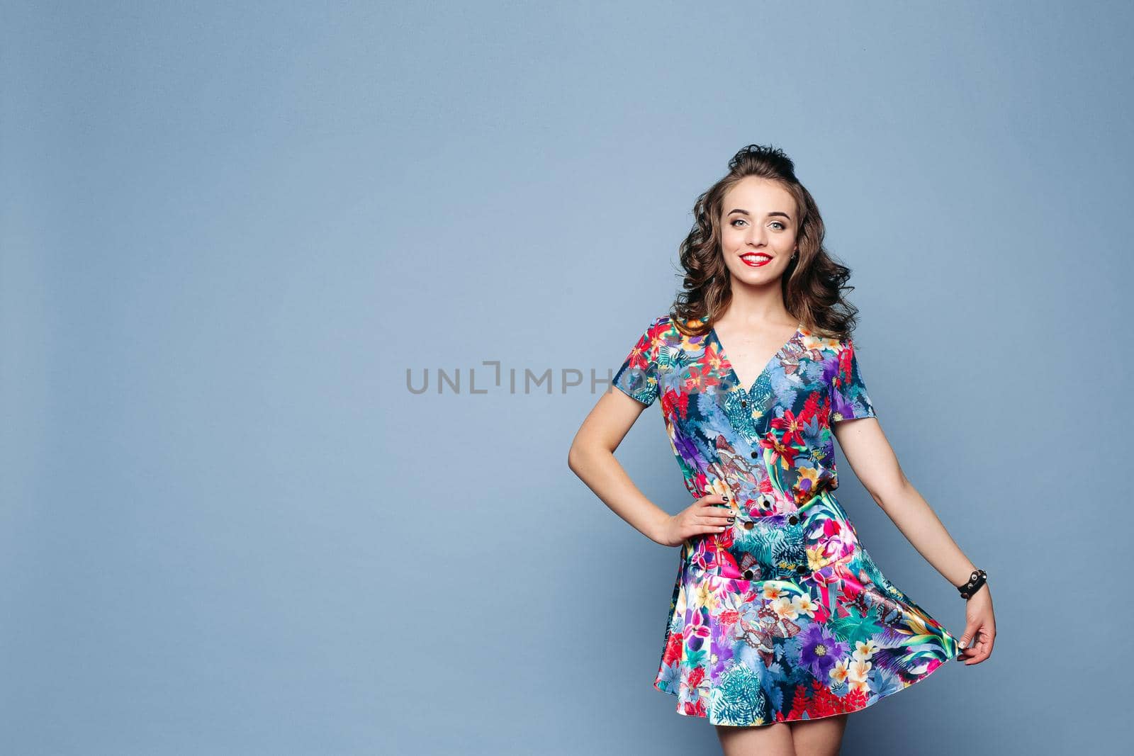 Portrait of attractive happy brunette lady with wavy hair in lovely floral dress holding skirt with arm and smiling at camera. Cutout on blue background. Studio shot.