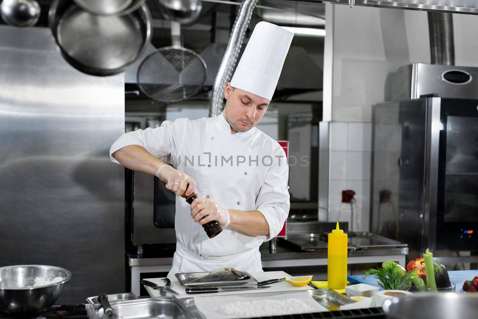 Professional chef in white uniform salts fish. Wooden salt grinder